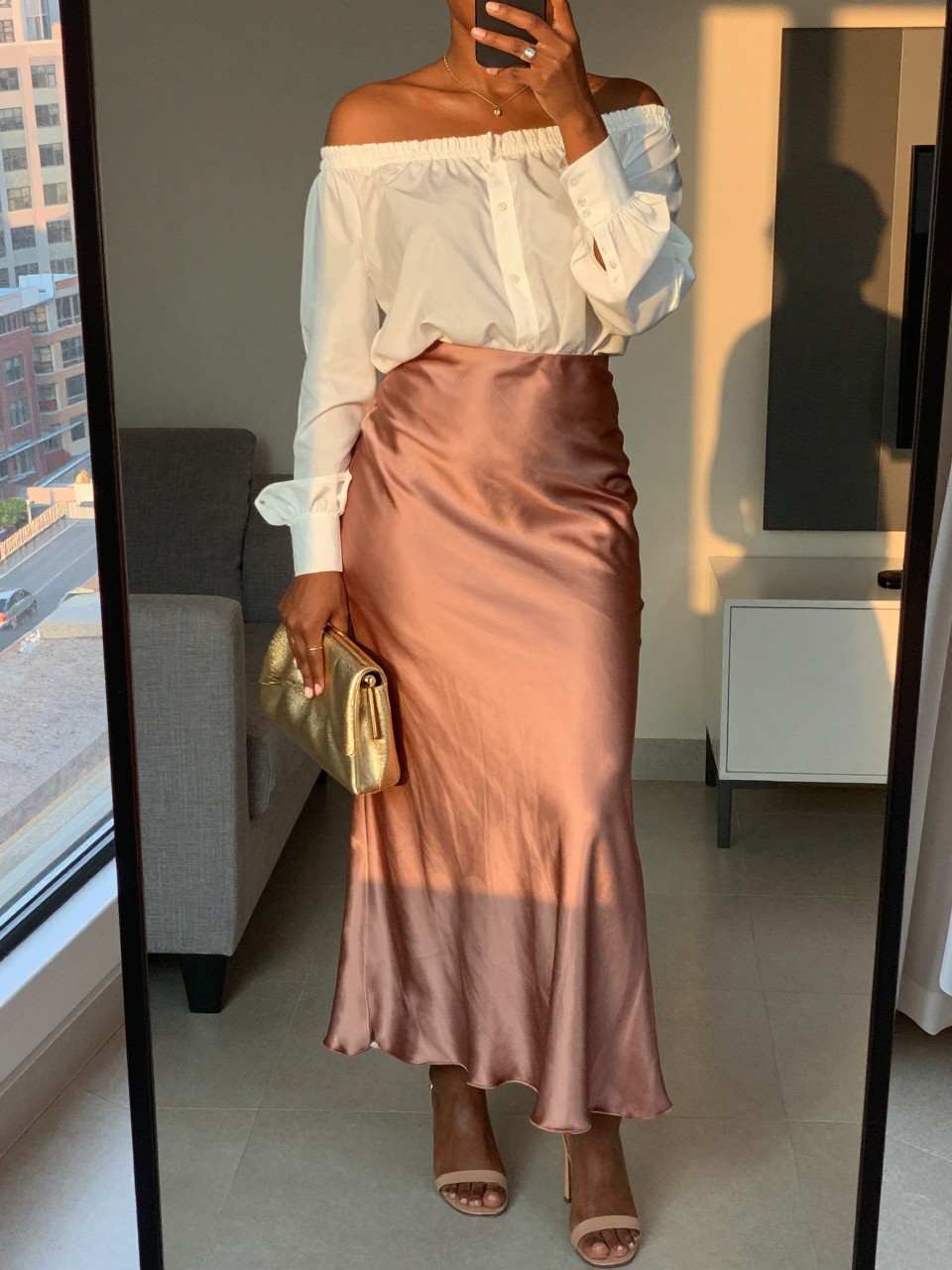 iPhone mirror selfie of a black woman wearing a white off-the-shoulder blouse tucked into a rose gold satin maxi skirt with nude block-heel sandals, holding a gold metallic clutch at her side, cropped at NECK so face doesn't show, in a city apartment, evening golden hour, iPhone photo quality.