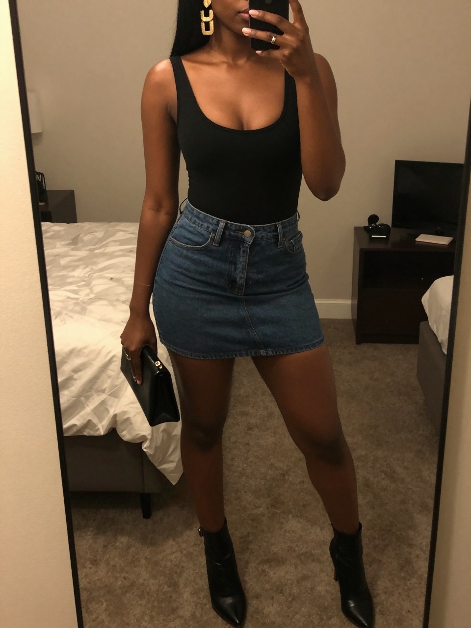 mirror selfie of a black woman wearing a black scoop-neck bodysuit tucked into a dark-wash denim mini skirt with black heeled ankle boots and bold gold statement earrings, holding a small black clutch, standing in a confident evening pose, cropped at NECK so face doesn't show, in a feminine-themed bedroom, warm moody lighting, iPhone photo quality.