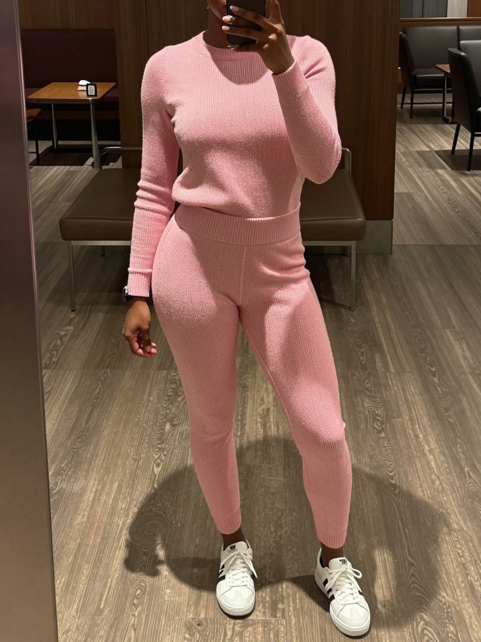 mirror selfie of a black woman in a pink knit set and white sneakers, standing in an airport or hotel lobby, cropped at NECK so face doesn't show, in a city apartment, warm moody lighting, iPhone photo quality.