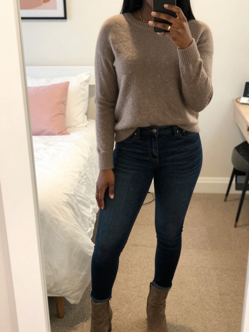 mirror selfie of a Black woman wearing a taupe sweater with dark wash jeans and boots, standing with her weight shifted and phone at her side, cropped at NECK so face doesn't show, in a bright feminine-themed bedroom, warm moody lighting, iPhone photo quality.