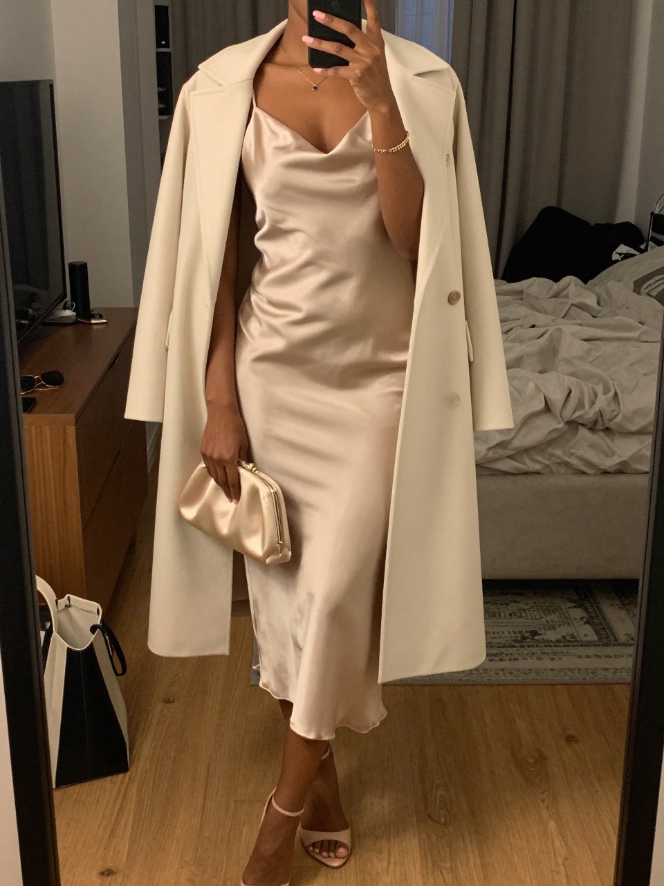 mirror selfie of a black woman wearing a champagne satin slip midi dress with a cream lightweight duster coat, nude strappy heels, a delicate gold bracelet, and a champagne satin small clutch, holding a phone in front of a mirror, cropped at NECK so face doesn't show, with a natural casual selfie pose, in a city apartment, warm moody lighting, including small lived-in details like a tote bag on the floor, sunglasses on a dresser, or soft bedding in the background, latest iPhone photo quality.