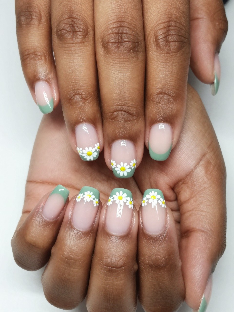 Close-up, overhead view of black feminine hands with a French manicure. The tips are a soft sage green. A delicate line of tiny white daisies with yellow centers is painted right at the border of the green tip. The look is clean, fresh, and subtly detailed.