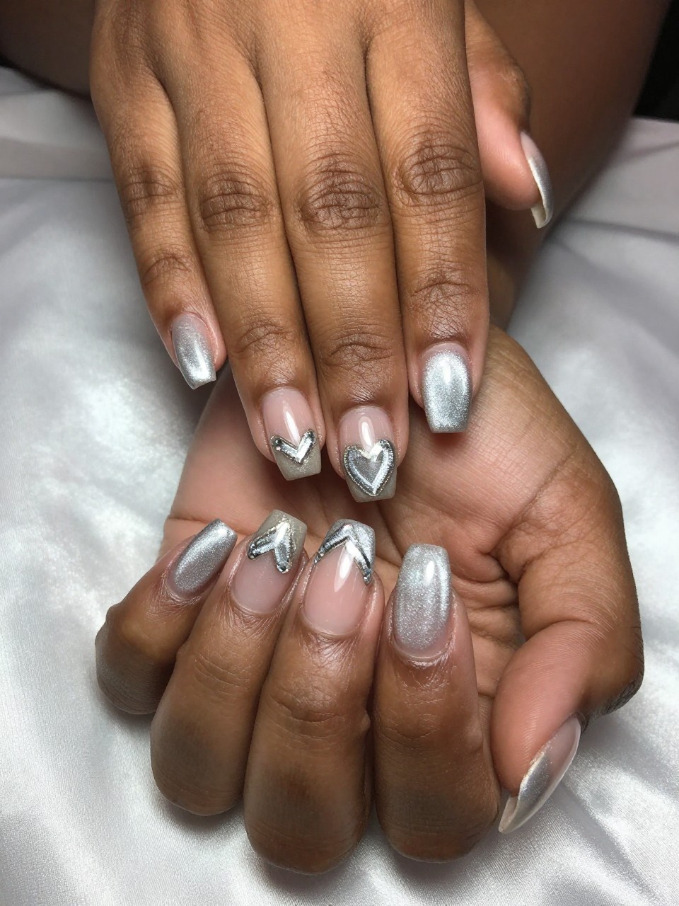 Close-up of black feminine hands with a French manicure where the tips are made of shimmering silver cat eye polish, concentrated into a heart shape. Hands rest on a silk pillow, with the heart tips catching the light brilliantly.