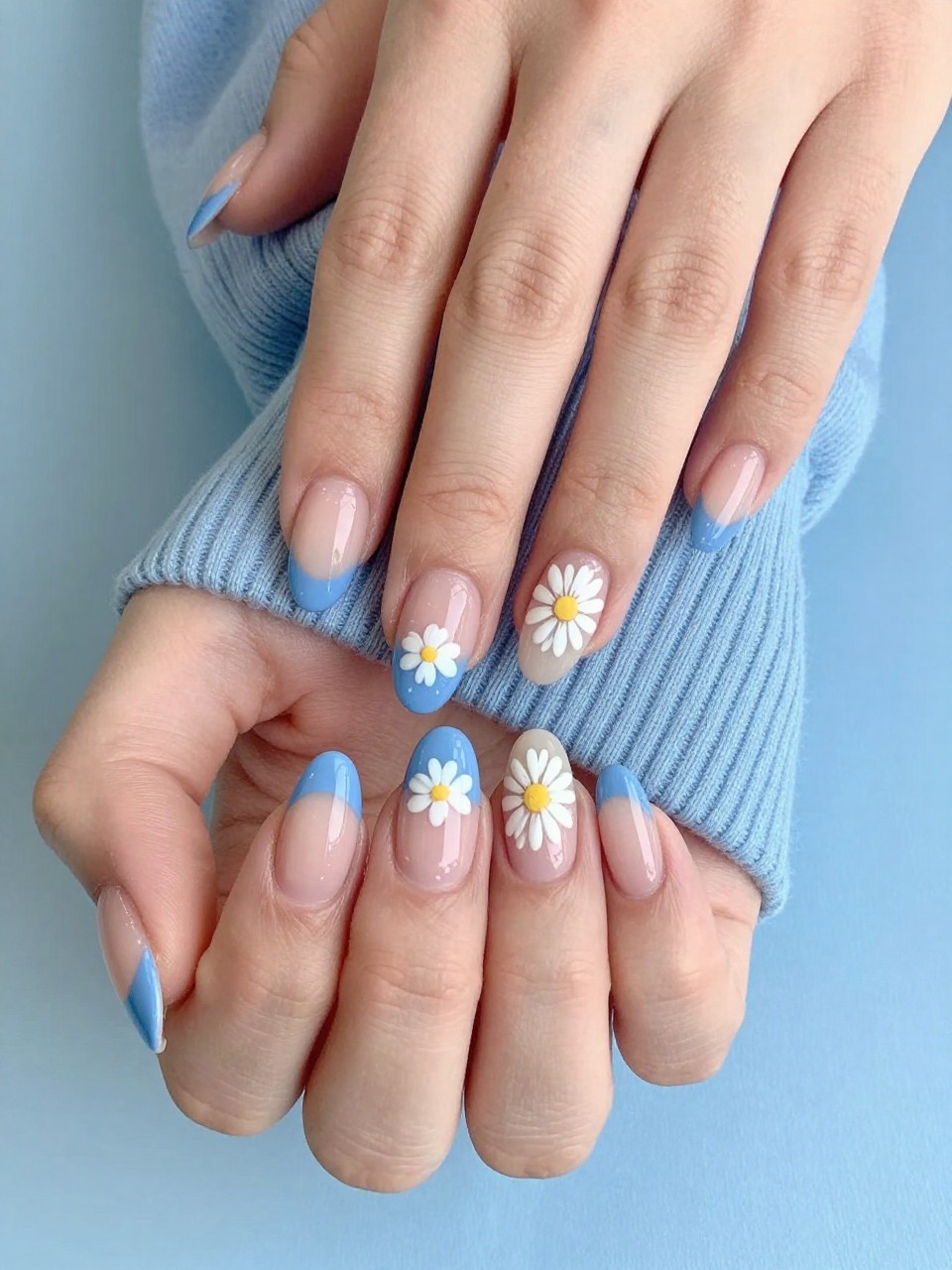Overhead view of hands with a soft blue French tip. On the line where the blue meets the nude nail, a pair of micro daisies are painted on each nail. The hands are resting on a light blue sweater, looking fresh and happy.