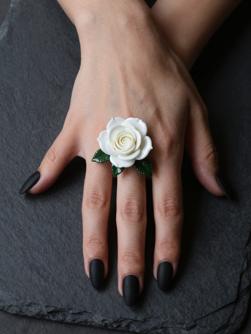 Overhead, top-down view of feminine black hands with almond nails resting on a dark slate surface. The nails are a flat matte black. While one ring finger design features a single, intricately shaded glossy white rose with green leaves. Moody, directional lighting highlights the textural contrast.