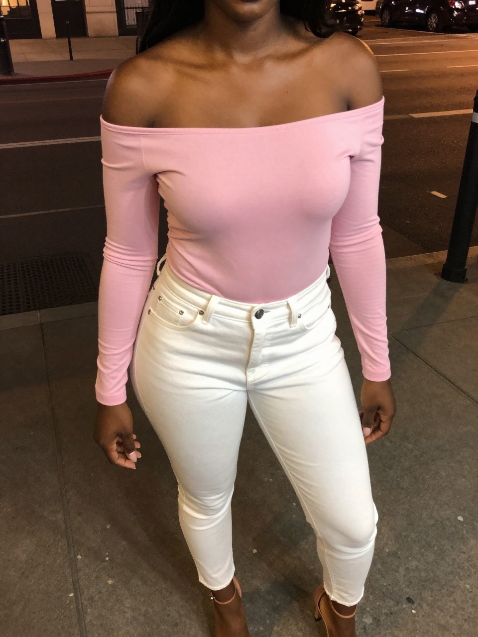 selfie of a black woman in a pink off-shoulder top and white jeans with heels, standing on a city street or boardwalk, cropped at NECK so face doesn't show, in a city apartment, warm moody lighting, iPhone photo quality.