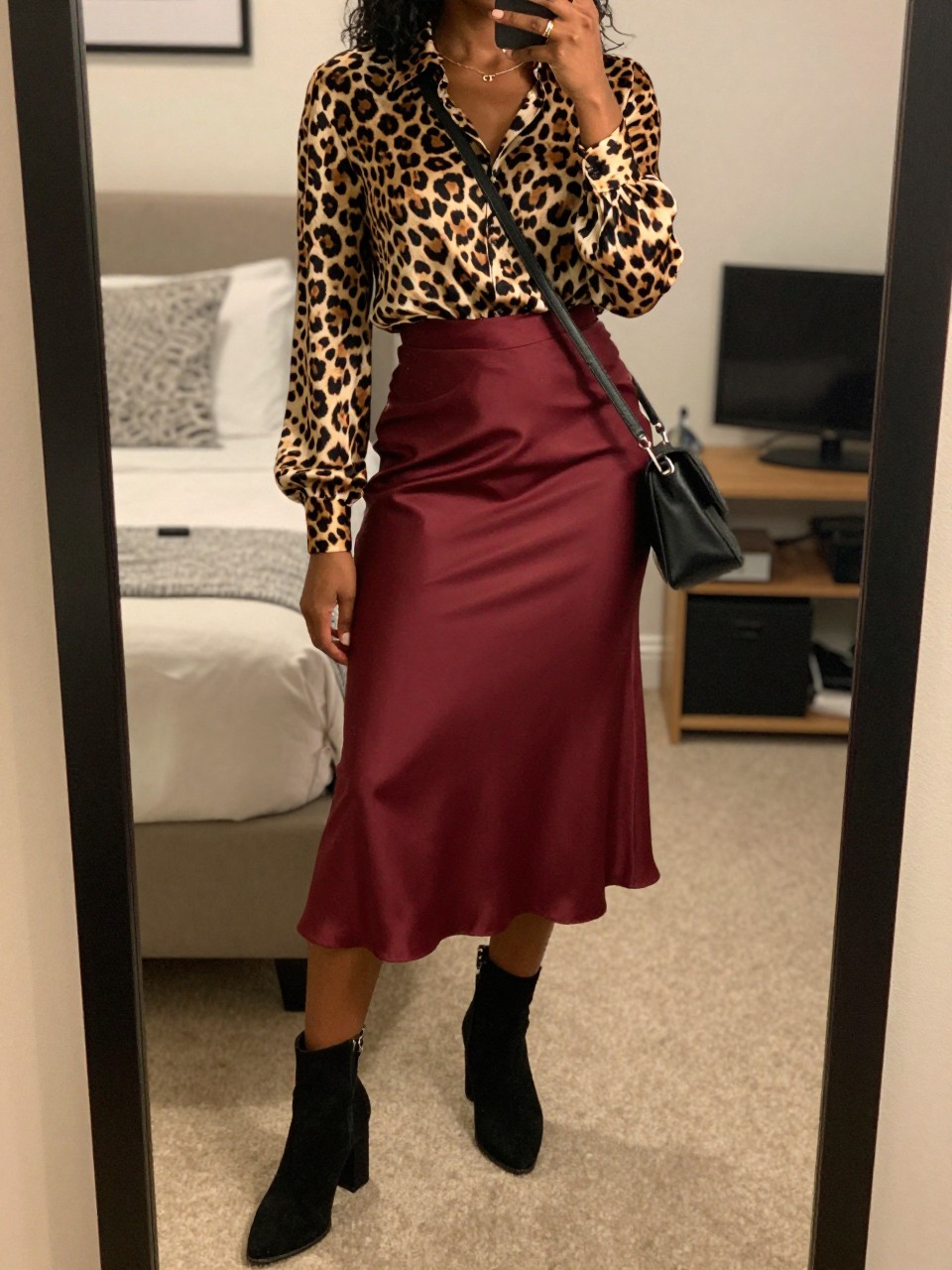 mirror selfie of a black woman wearing a leopard print silk blouse tucked into a burgundy red satin midi skirt with black suede block-heel ankle boots and a black crossbody bag, standing with her weight on one leg, cropped at NECK so face doesn't show, in a feminine-themed bedroom, warm moody lighting, iPhone photo quality.