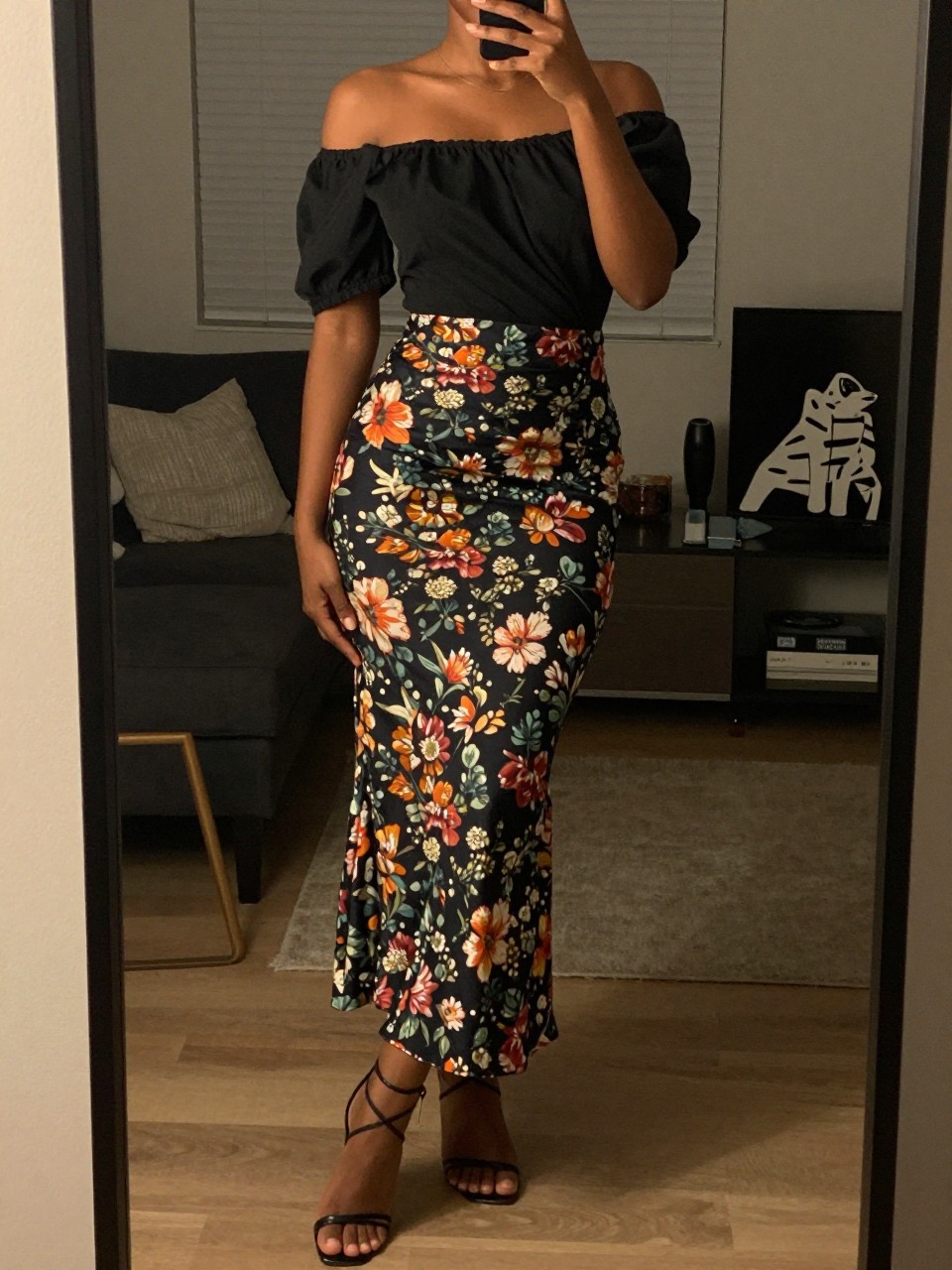 iPhone mirror selfie of a black woman in an off-the-shoulder top and a jewel-toned floral maxi skirt with strappy heels, posing with one foot forward, cropped at NECK so face doesn't show, in a city apartment, warm moody lighting, iPhone photo quality.