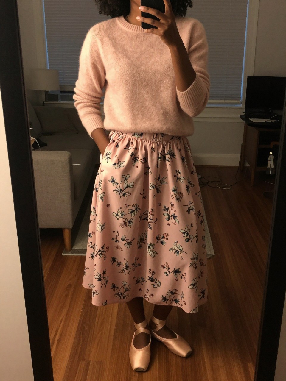 iPhone mirror selfie of a black woman in a blush pink cashmere sweater and a pink floral skirt with ballet flats, standing with one hand in her pocket, cropped at NECK so face doesn't show, in a city apartment, warm moody lighting, iPhone photo quality.