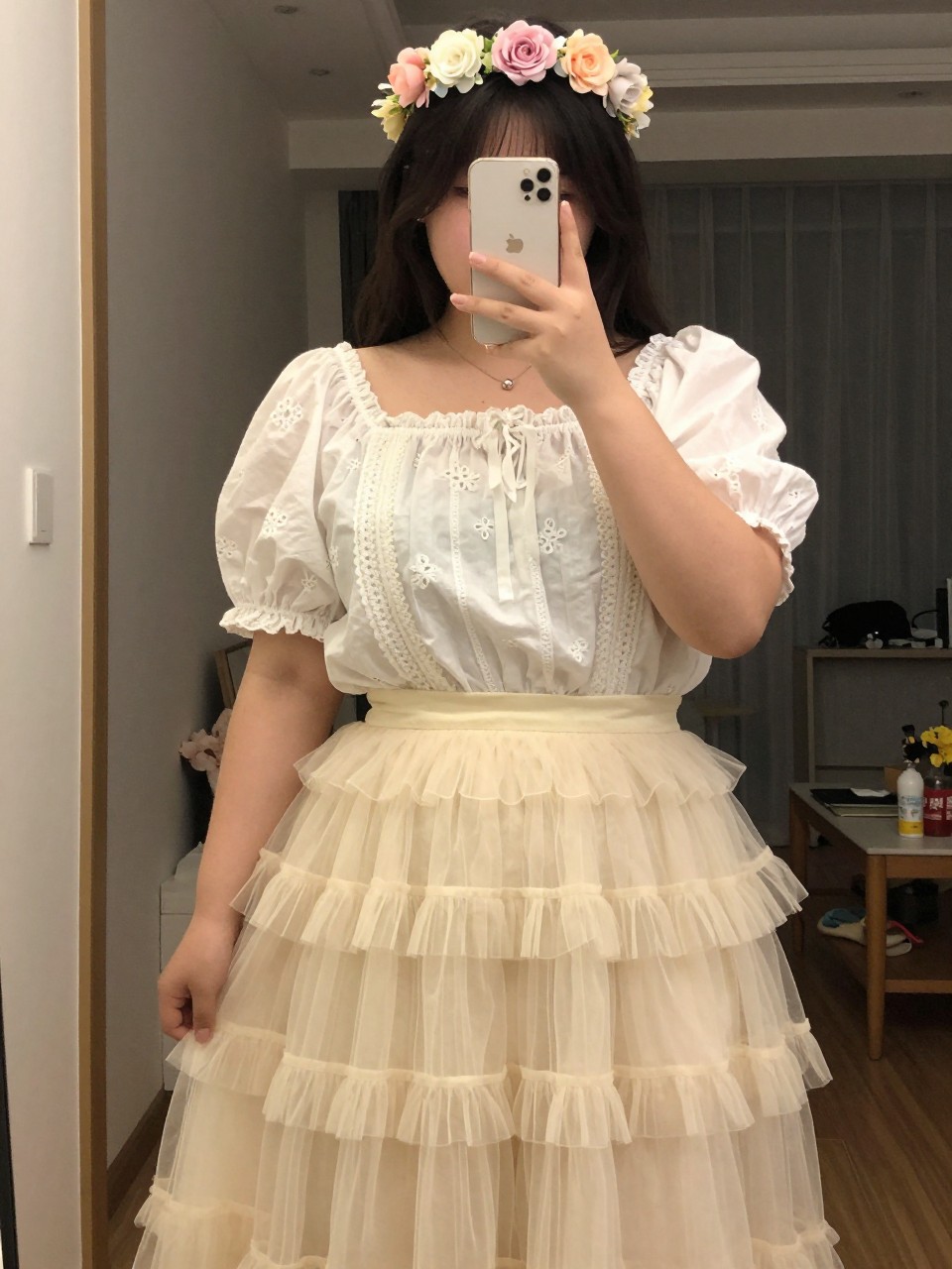 mirror selfie of a curvy woman wearing a white embroidered puff-sleeve blouse with a layered cream tulle fairy skirt and a pastel flower crown, one hand holding her phone at chest level, cropped at NECK so face doesn't show, in a city apartment, warm moody lighting, iPhone photo quality.