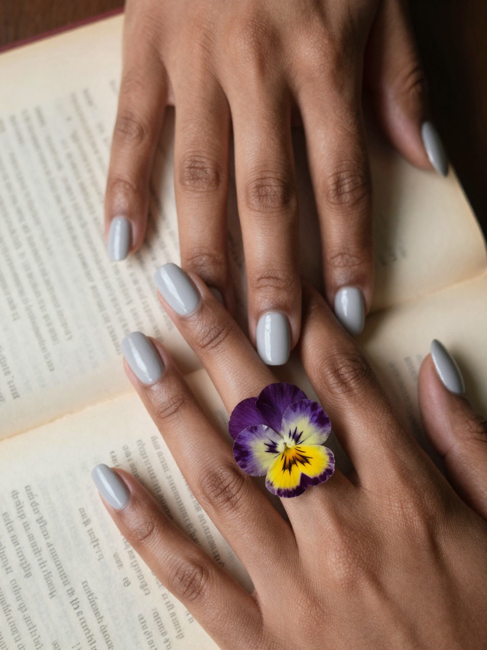 Macro, top-down view of black feminine hands over an open, old book. The nails are a matte pale grey. One ring finger features a real, pressed purple and yellow pansy perfectly sealed under glass-like glossy gel. The light is soft, focused, and slightly vintage.