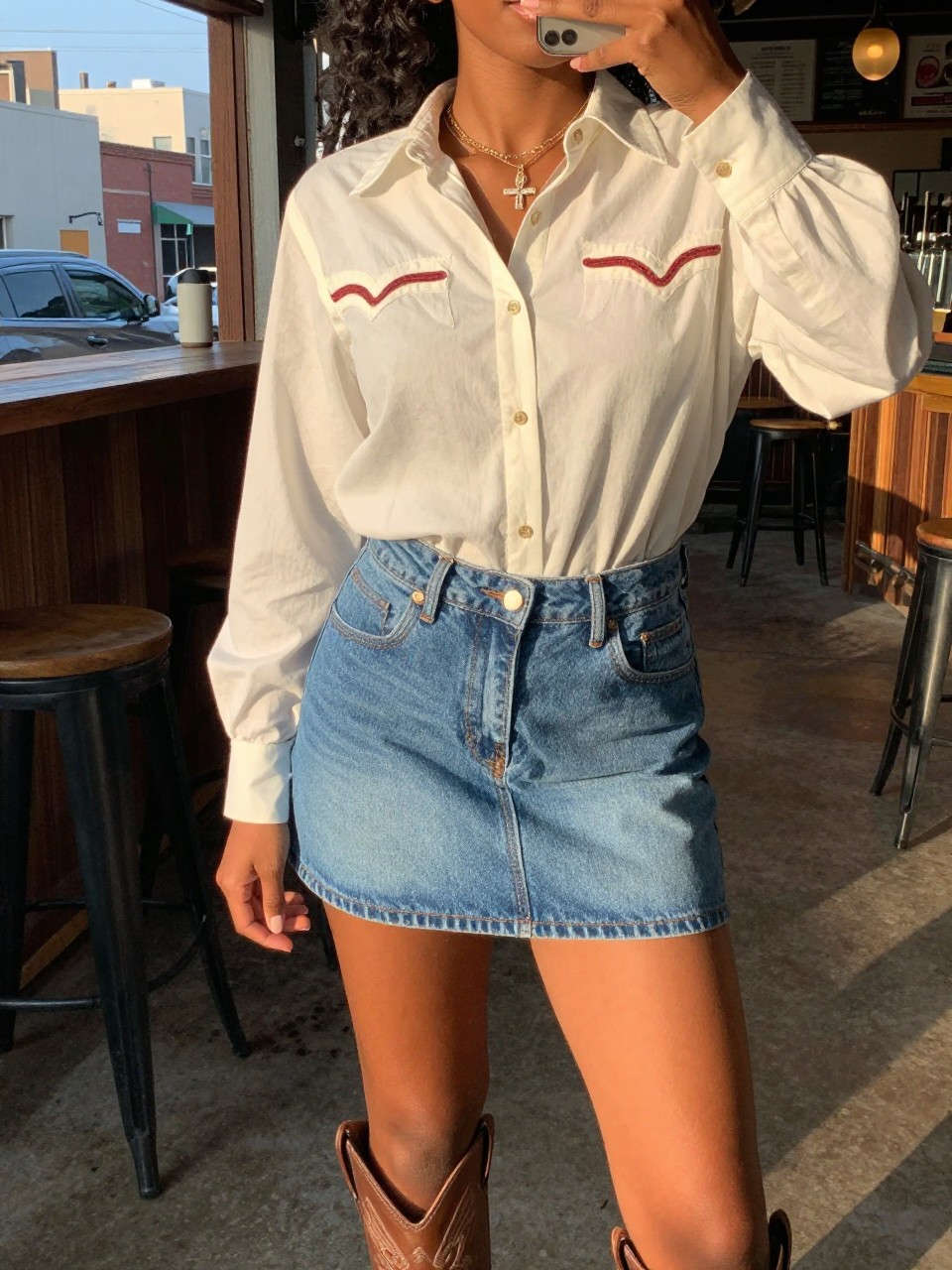 selfie of a black woman in a denim skirt and Western blouse with cowboy boots, standing at a rustic venue or bar, cropped at NECK so face doesn't show, in a city apartment, evening golden hour, iPhone photo quality.