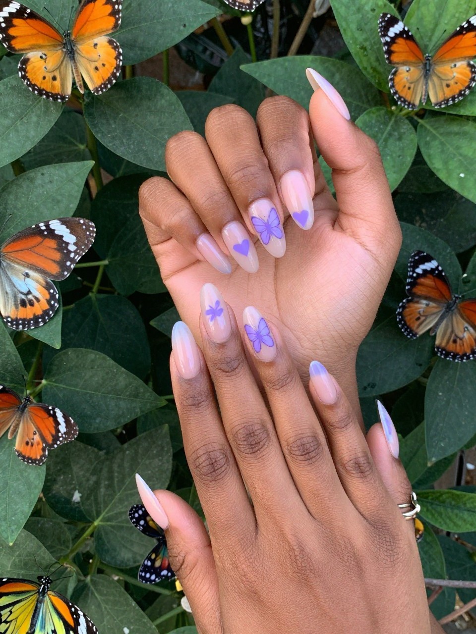 Bird's-eye view of black feminine hands with almond nails posed near live butterflies in a garden. The nails have a sheer base with simple lavender butterfly silhouettes and small hearts. The mood is whimsical and light.