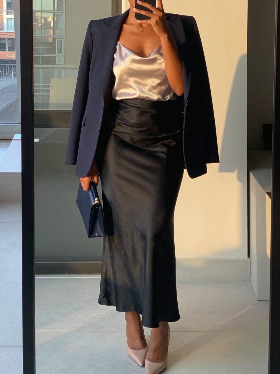 mirror selfie of a black woman wearing a champagne silk camisole tucked into a black satin maxi skirt with a structured navy blazer worn open and nude pointed heels, holding a small navy clutch, standing in a polished professional pose, cropped at NECK so face doesn't show, in a city apartment, evening golden hour, iPhone photo quality.