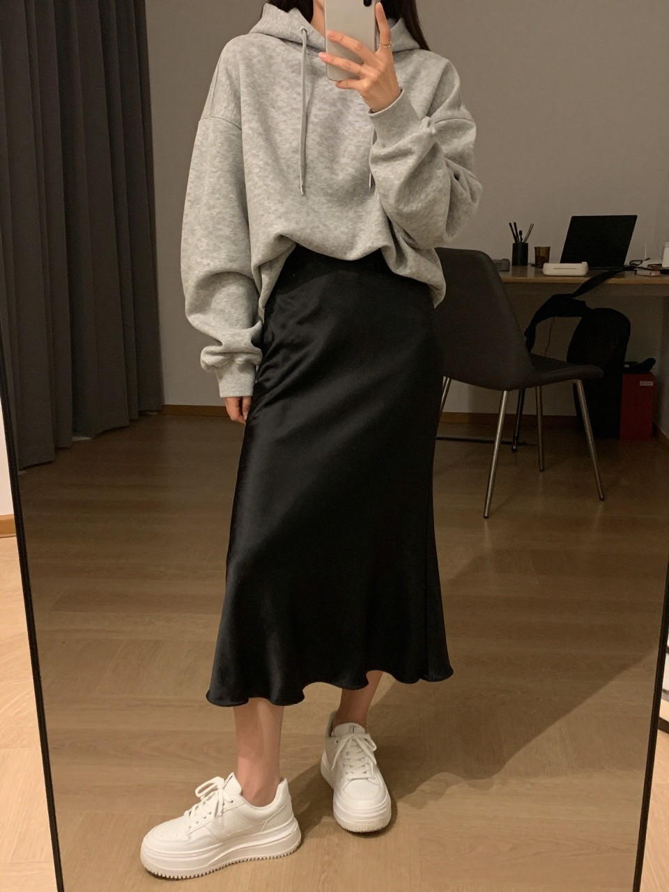 iPhone mirror selfie of a woman wearing an oversized gray hoodie half-tucked into a black satin midi skirt with chunky white platform sneakers, one hand holding her phone low by her thigh, cropped at NECK so face doesn't show, in a city apartment, warm moody lighting, iPhone photo quality.