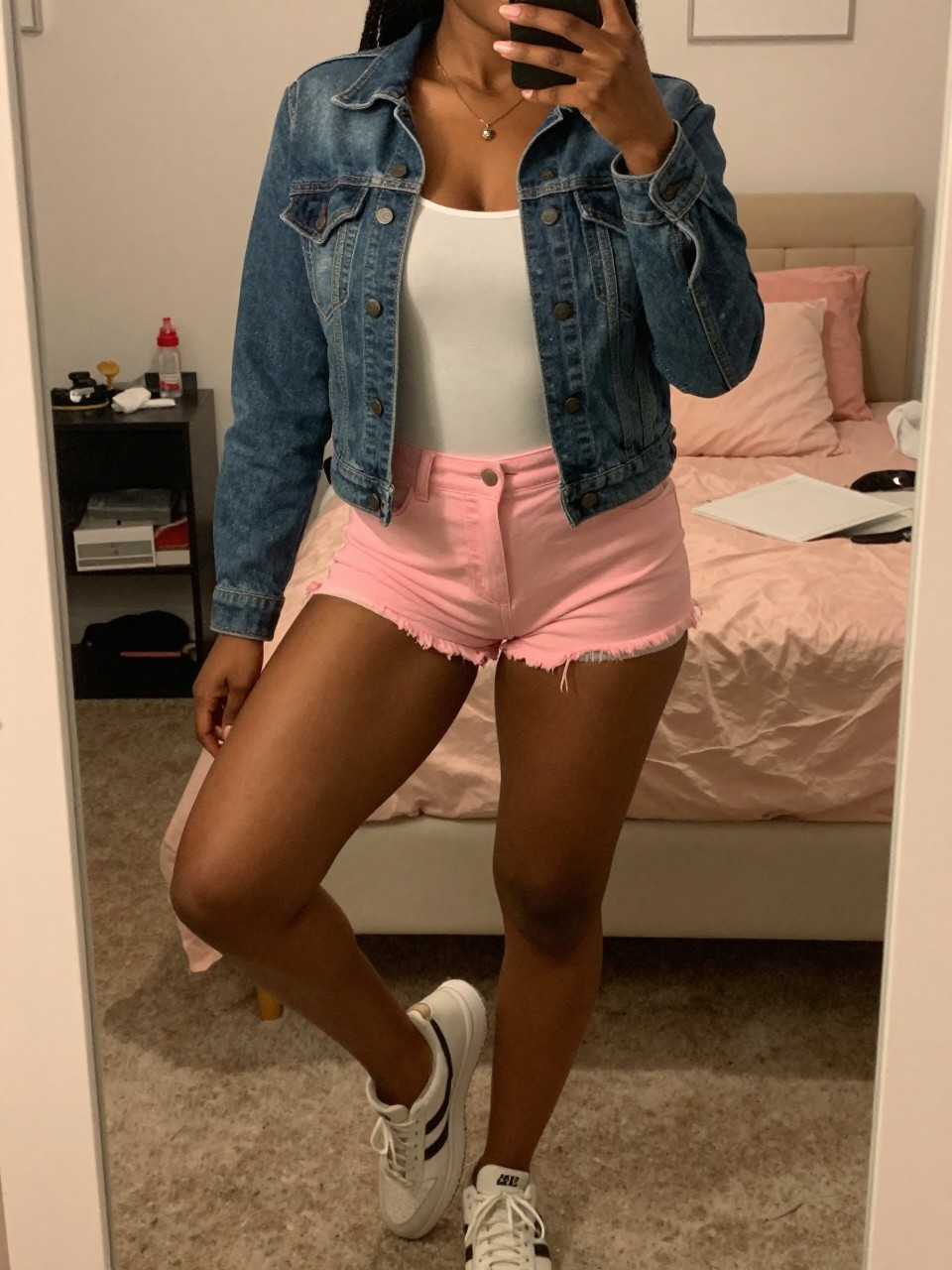 mirror selfie of a black woman in pink cutoff shorts and a denim jacket over a white tank with sneakers, standing with weight on back leg, cropped at NECK so face doesn't show, in a feminine-themed bedroom, warm moody lighting, iPhone photo quality.