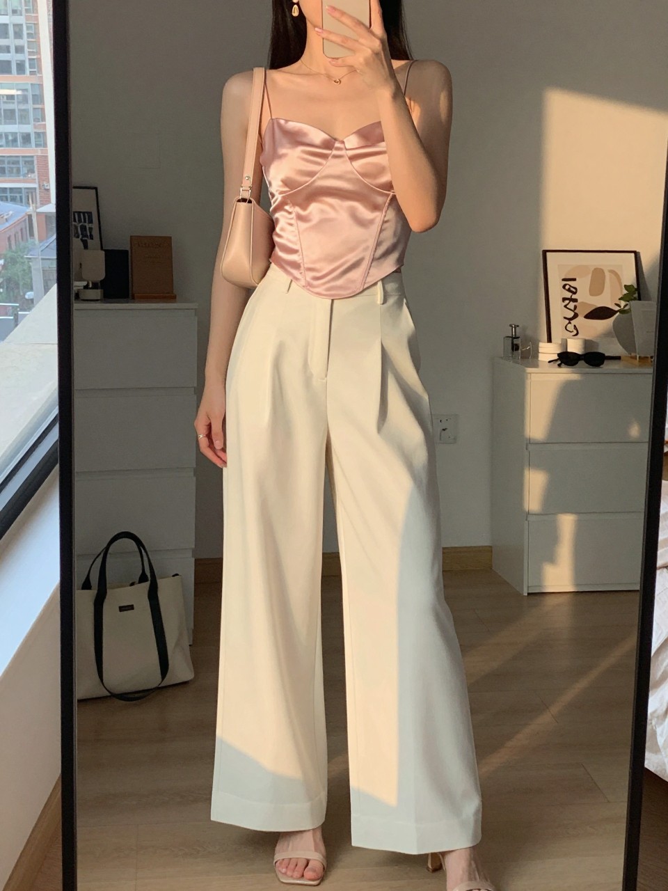 mirror selfie of a woman wearing ivory wide-leg trousers with a pale pink satin corset-style top, nude heeled sandals, a blush small shoulder bag, and simple gold drop earrings, holding a phone in front of a mirror, cropped at NECK so face doesn't show, with a natural casual selfie pose, in a city apartment, evening golden hour, including small lived-in details like a tote bag on the floor, sunglasses on a dresser, or soft bedding in the background, latest iPhone photo quality.