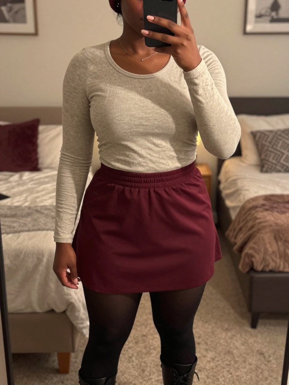 mirror selfie of a black curvy woman wearing a heathered oatmeal thermal tucked into a burgundy tennis skirt with black tights and combat boots, one hand adjusting her beanie, cropped at NECK so face doesn't show, in a feminine-themed bedroom, warm moody lighting, iPhone photo quality.