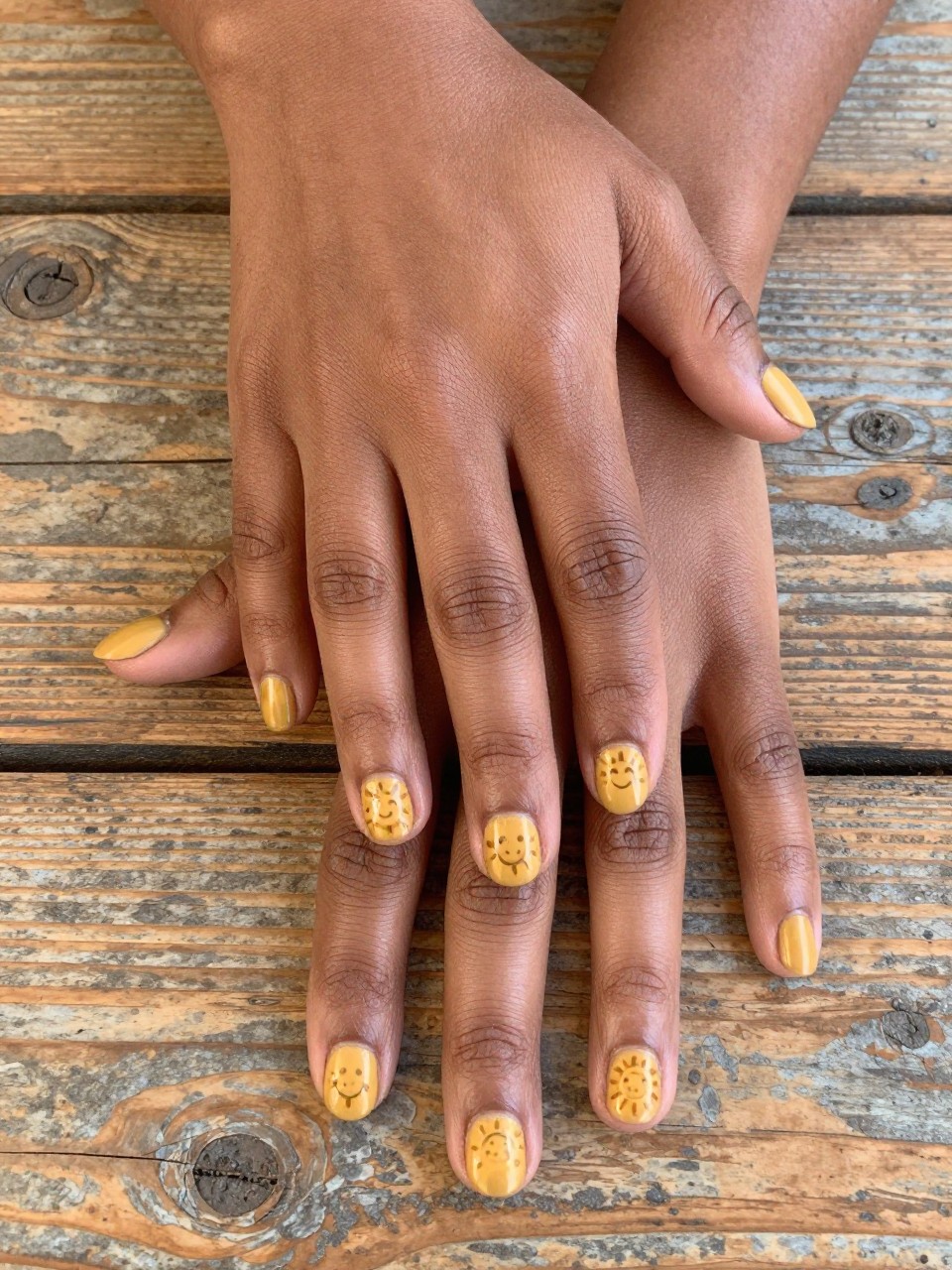 Overhead shot of black feminine hands with rounded nails on a rustic wooden table. The nails are a matte goldenrod yellow. Each nail features a simple, charming sun face with a smile and rays. The look is folksy, happy, and full of personality.