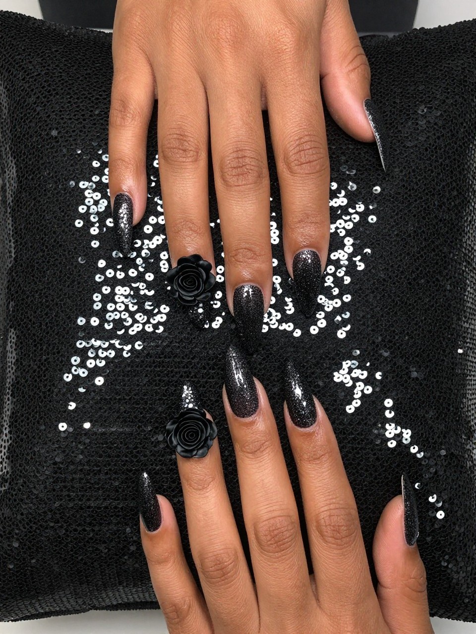Top-down view of black feminine hands with coffin nails resting on a sequined black pillow. The nails have a sparkling black glitter gradient. each nail design features a single, intricate black rose decal placed where the glitter is most dense. The mood is luxe and edgy.