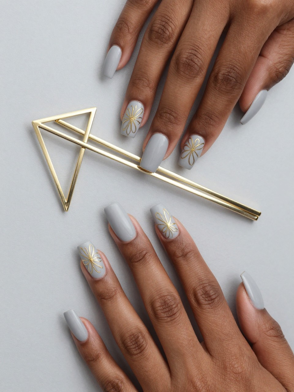 Overhead shot of black feminine hands with long, squared nails next to an art deco geometric hairpin. The nails are a matte pale grey. Precise gold lines form elegant, angular floral patterns. The look is modern, luxurious, and structured.