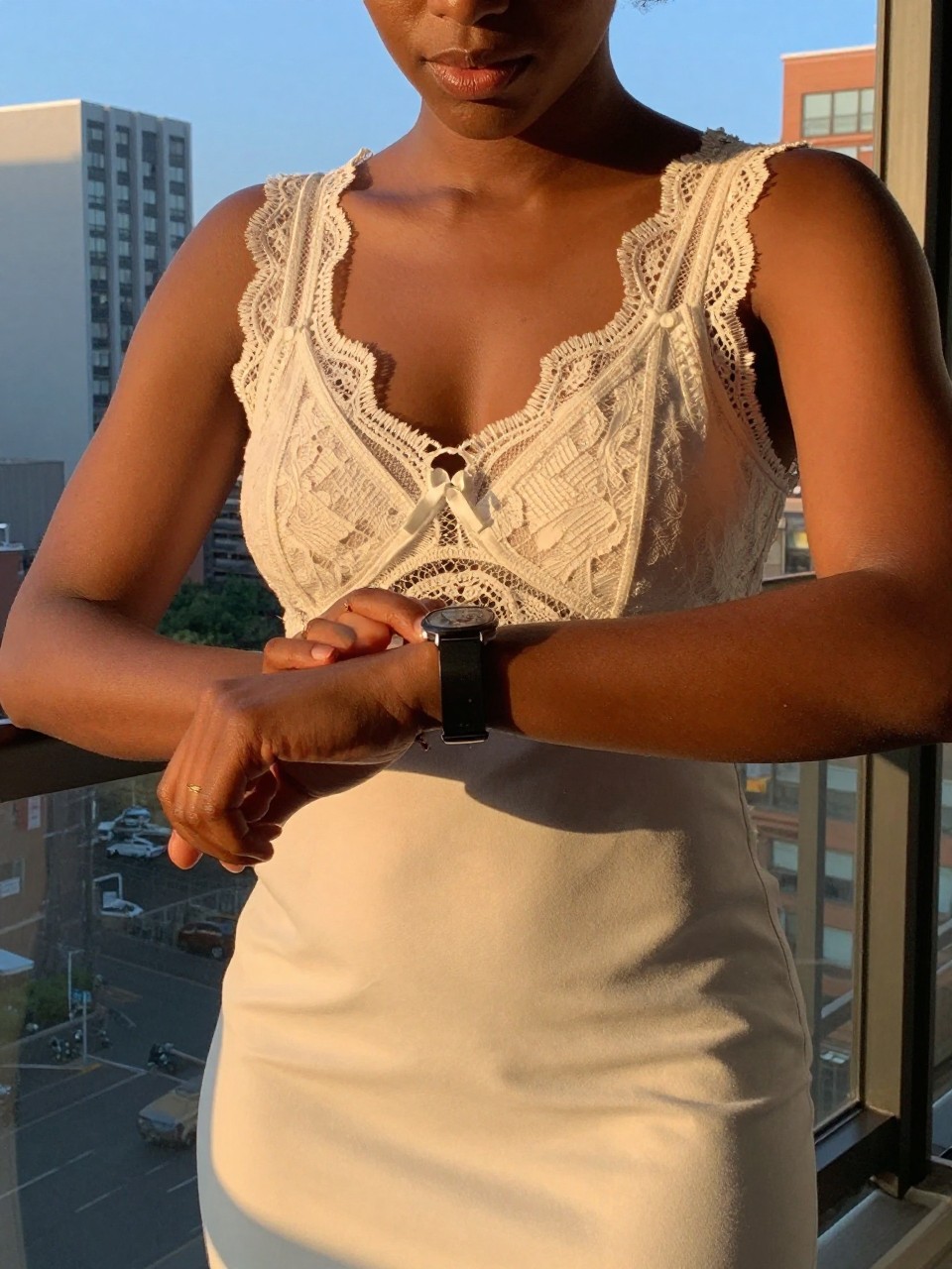 selfie of a black woman in a lace-trimmed dress, checking her bracelet watch, cropped at NECK so face doesn't show, in a city apartment, evening golden hour, iPhone photo quality.