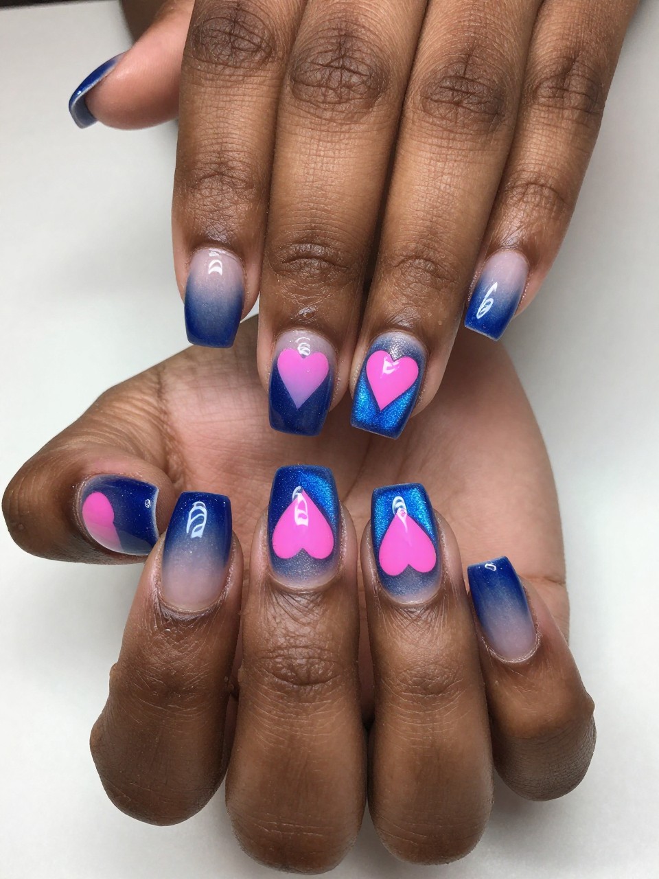 Overhead view of black feminine hands with a magnetic blue cat eye French tip. A heart-shaped hole in the center of each tip reveals a bright pink underlayer. The look is fresh, structured, and colorful.