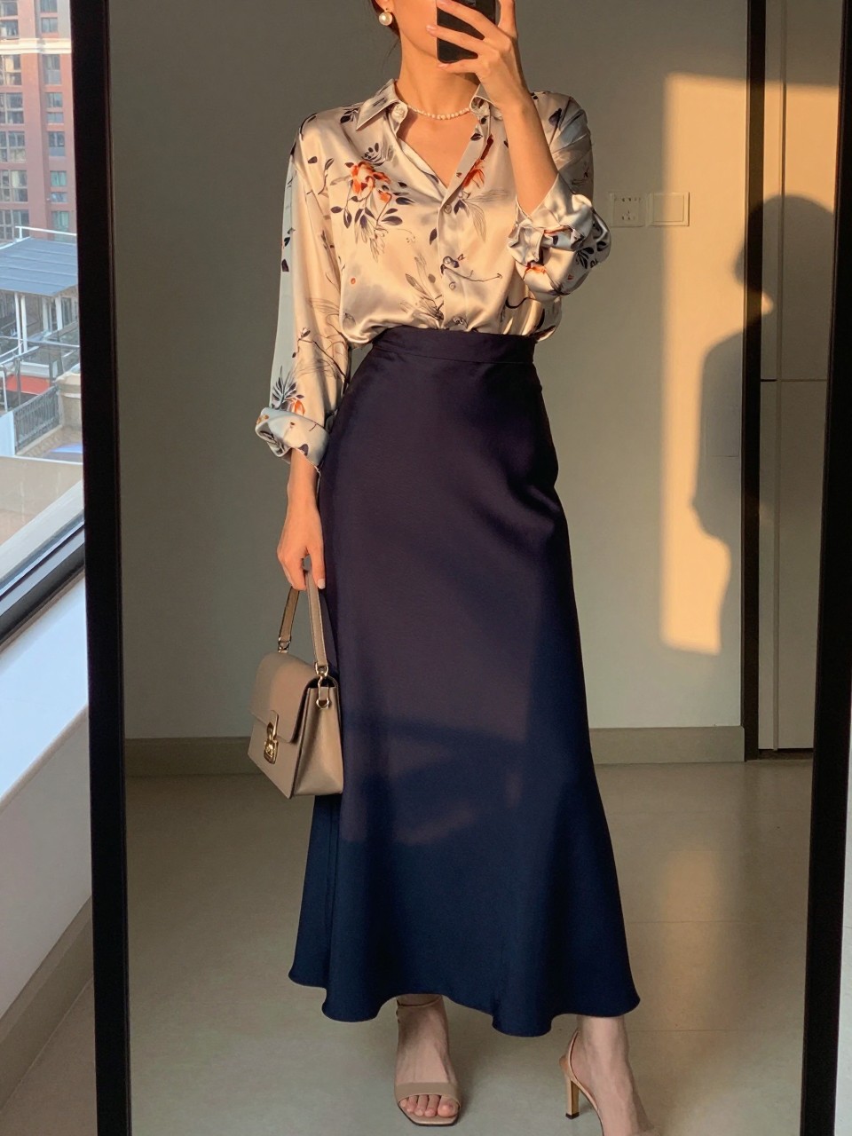 mirror selfie of a woman wearing a soft floral print silk blouse tucked into a navy blue maxi skirt with nude heeled sandals and pearl stud earrings, holding a small taupe shoulder bag, standing in a romantic pose, cropped at NECK so face doesn't show, in a city apartment, evening golden hour, iPhone photo quality.