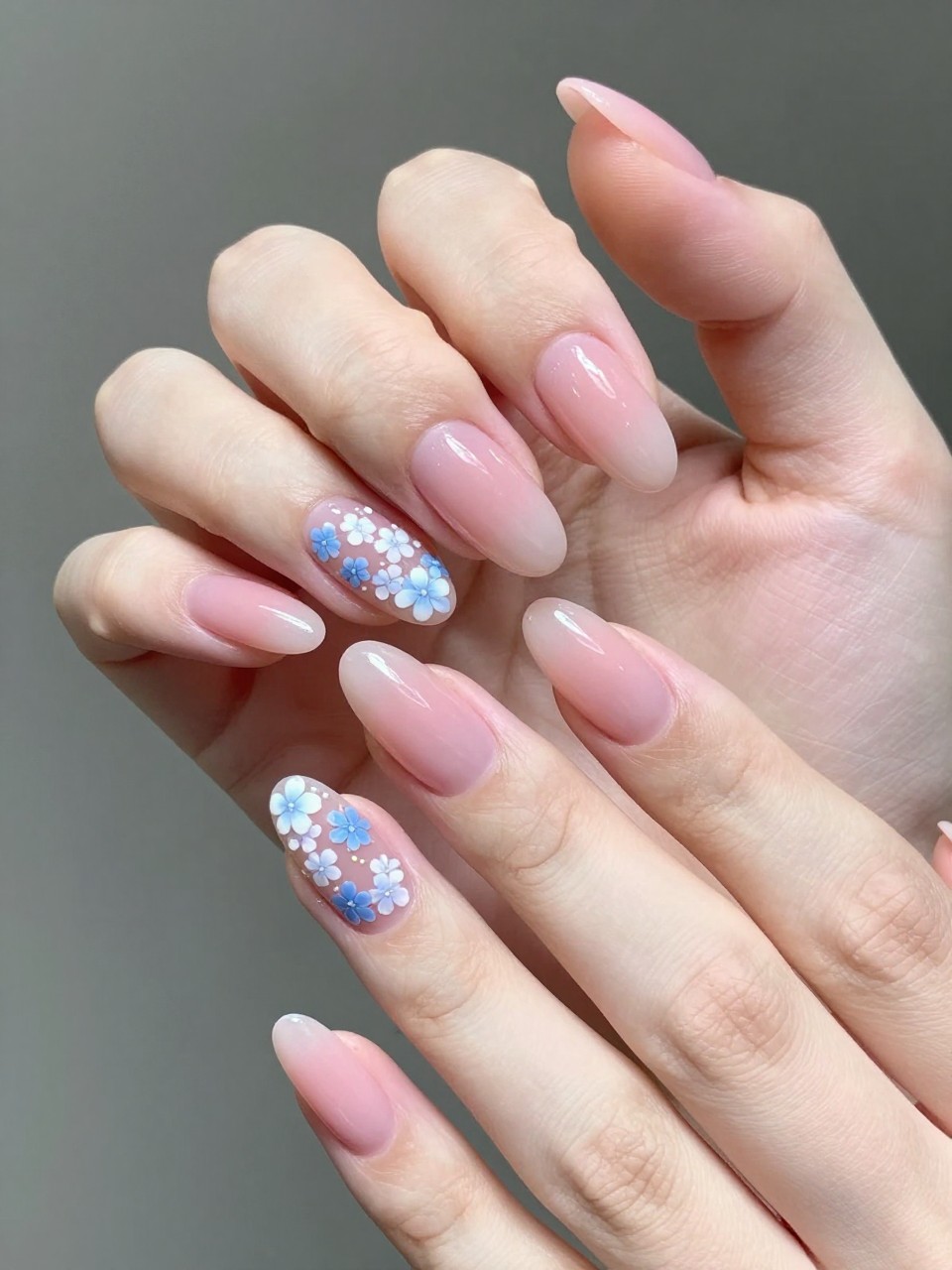 Angled view of hands with almond nails. The tips have a gentle pink ombré. A delicate line of tiny blue and white flowers is painted precisely on the ombré transition. The effect is soft, detailed, and beautifully blended.