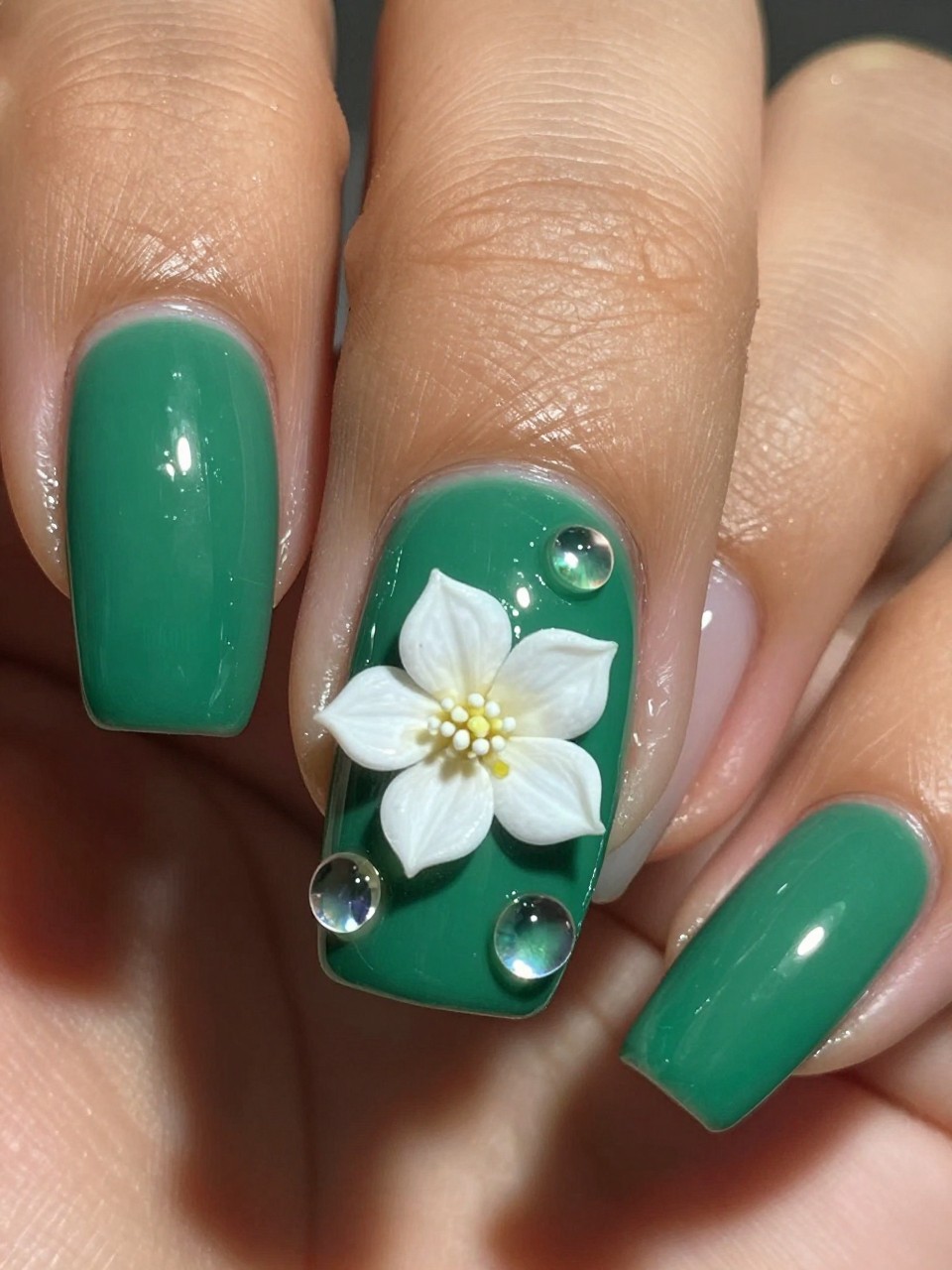 Macro, top-down view of a black feminine nail under bright light. The base is a glossy emerald green. The nail design features several clear, dimensional gel droplets are placed around a single, small white gardenia. The light catches the droplets, creating a wet, magical effect.