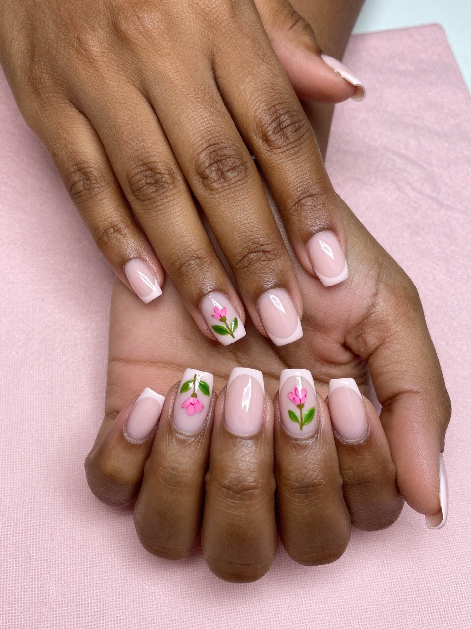 Close-up, bird's-eye view of black feminine hands with a classic French manicure. The tips are a soft blush pink. At the seam on each nail, one tiny pink flower bud with a single green leaf is painted. The hands rest on a pale pink linen napkin, looking clean and pretty.