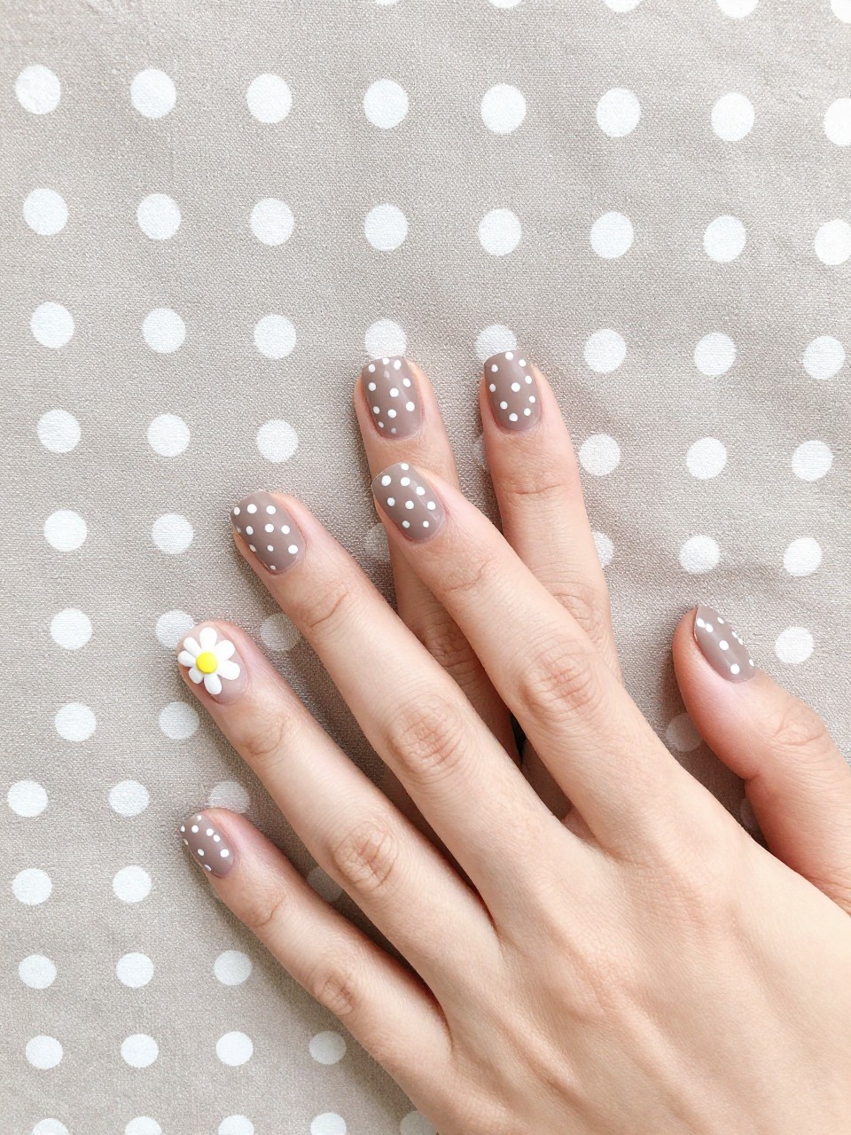 Bird's-eye view of hands with short, squared nails on a taupe and white polka dot fabric. The nails are matte taupe with white polka dots. The ring fingers feature a small white flower with a yellow center amidst the dots. The setting is bright and preppy.