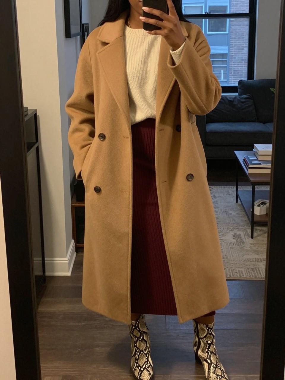mirror selfie of a black woman wearing a long camel coat over a cream sweater and maroon ribbed skirt with snake print boots, one hand holding her phone, cropped at NECK so face doesn't show, in a city apartment, warm moody lighting, iPhone photo quality.