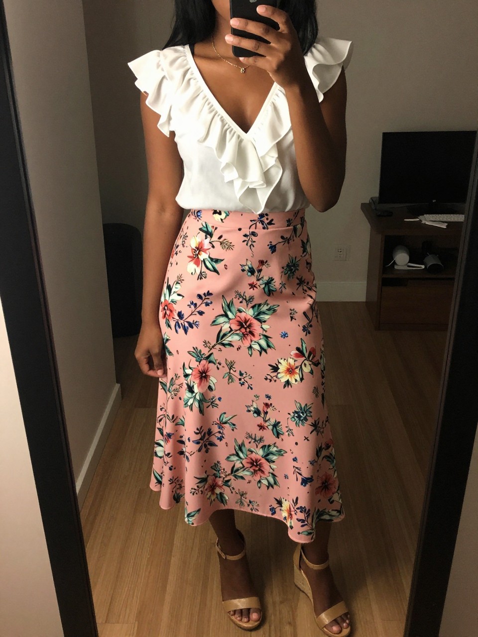 iPhone mirror selfie of a black woman in a ruffled white blouse and a pink floral skirt with wedge sandals, gently touching her hair, cropped at NECK so face doesn't show, in a city apartment, warm moody lighting, iPhone photo quality.