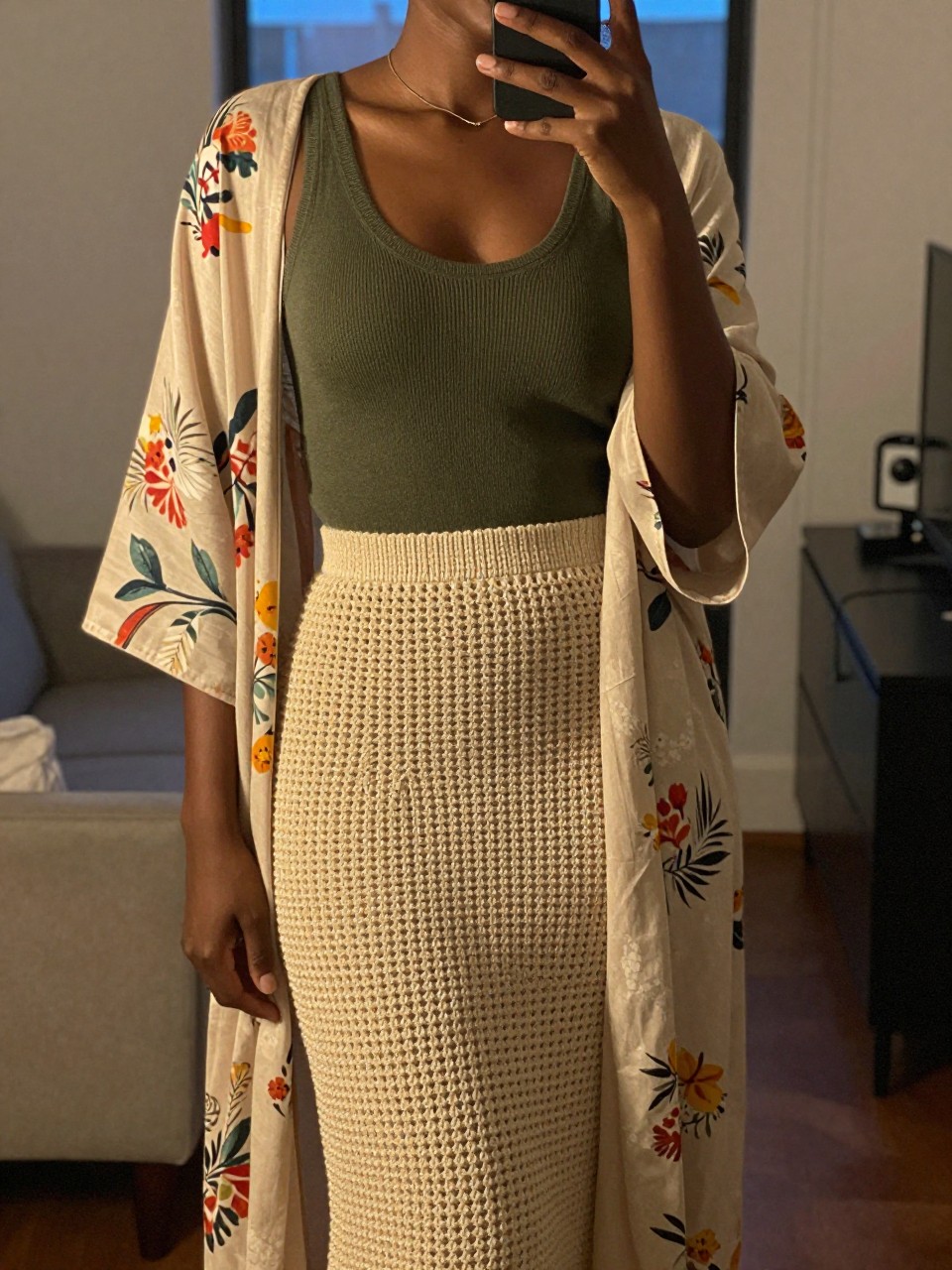 mirror selfie of a black woman wearing an olive green knit tank top with a natural crochet midi skirt and a lightweight printed floral kimono over it, one hand holding her phone at her side, cropped at NECK so face doesn't show, in a city apartment, warm moody lighting, iPhone photo quality.