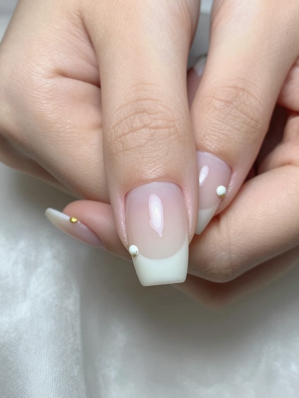 Macro, top-down view of nails under soft light. The base is sheer, milky white. Several tiny white buds are painted, each with a speck of gold at its stem. The hands rest on a silk cloth, with the gold details catching the light subtly.