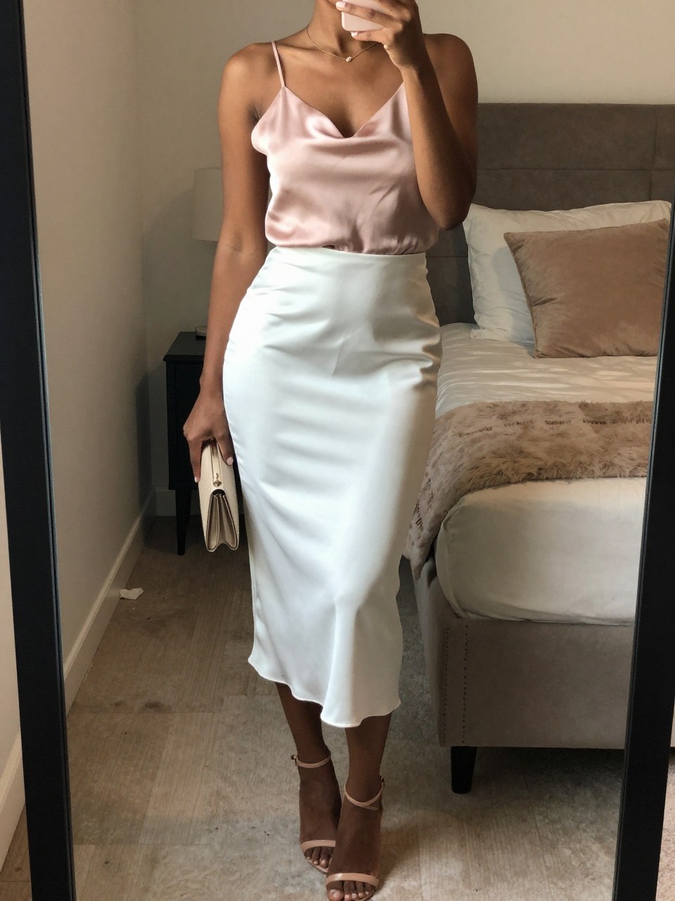 mirror selfie of a black woman wearing a blush pink silk camisole tucked into a white satin midi skirt with nude strappy heels, holding a small clutch, standing in a slight angle pose, cropped at NECK so face doesn't show, in a feminine-themed bedroom, warm moody lighting, iPhone photo quality.
