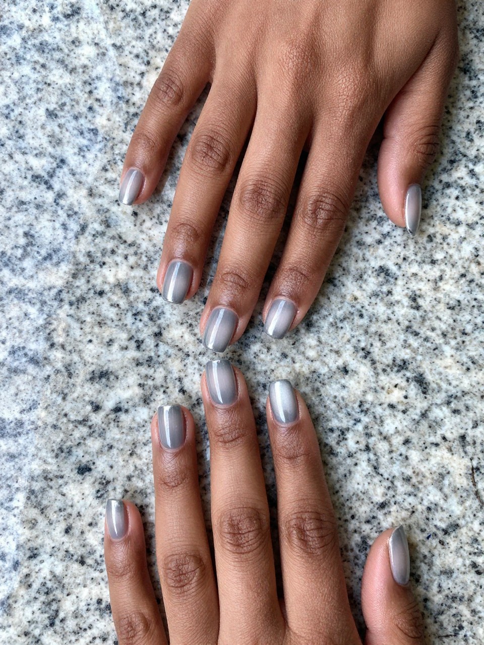 Overhead view of black feminine hands resting on a piece of cool, polished granite. The nails are a short square shape. The ombré transitions from a matte light grey at the cuticles to a glossy liquid silver at the free edge. The lighting is cool, bright, and modern.