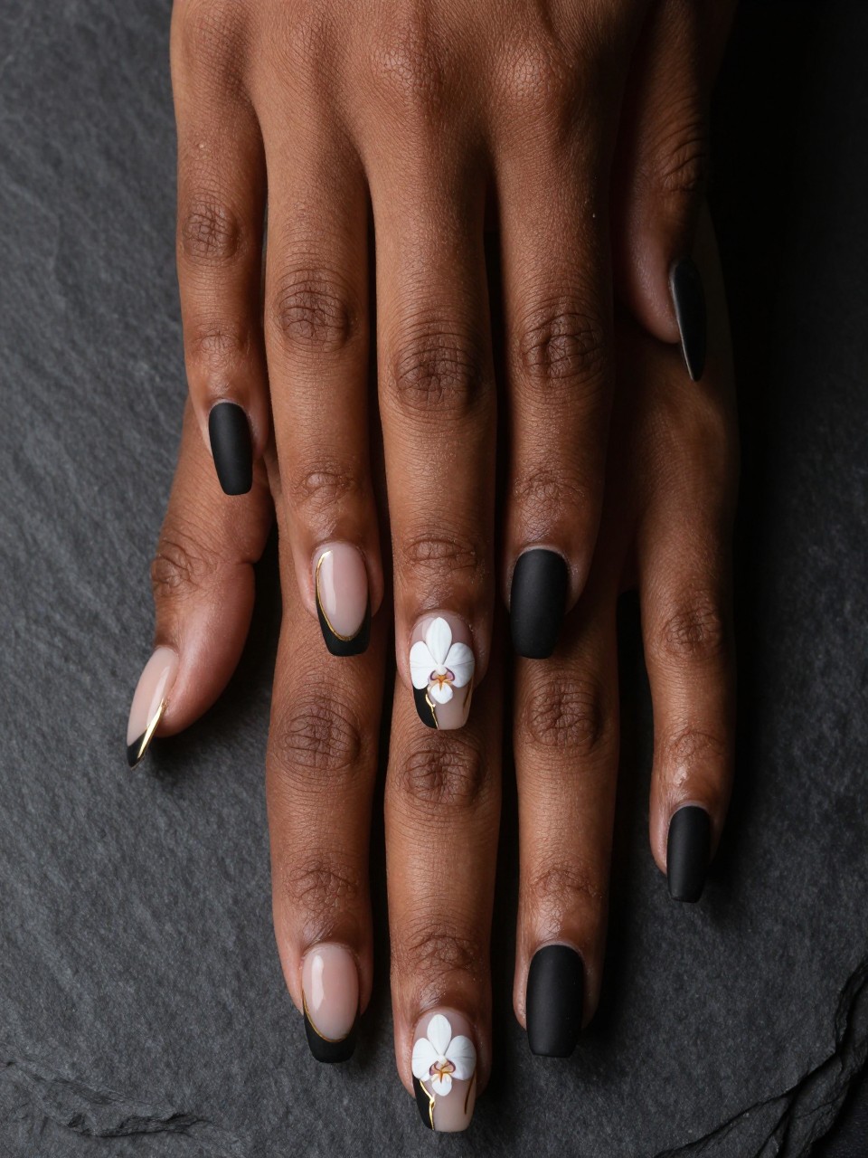 Top-down view of black feminine hands with coffin nails against a dark slate background. The nails have matte black French tips. on the nail design, there is a white orchid is painted on the nude base of the nails design with a fine gold line accenting its curves. The lighting is dramatic.