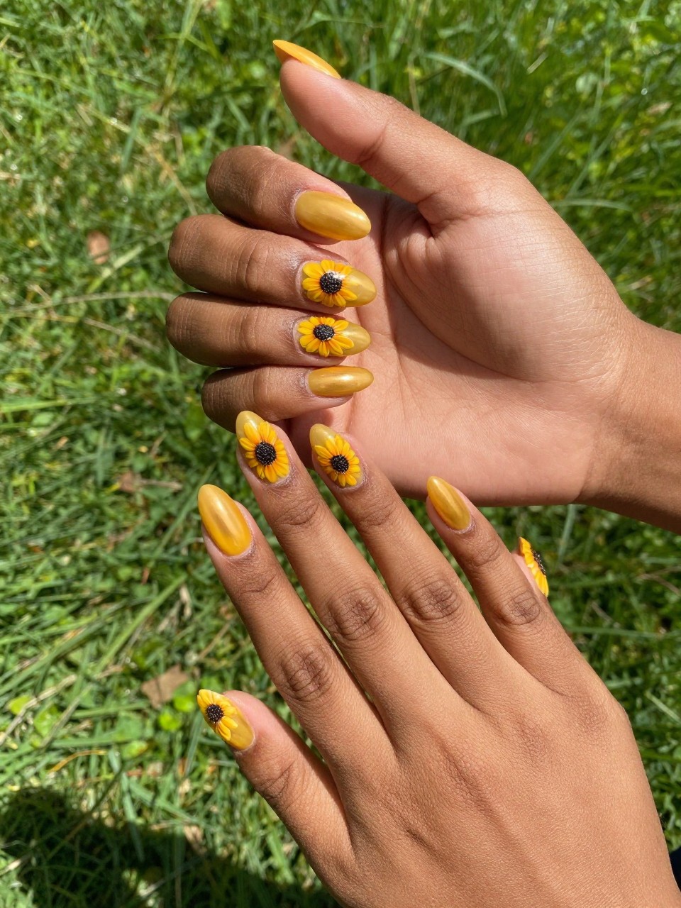 Bird's-eye view of black feminine hands with almond nails. The nails are a glossy golden yellow. Each nail design features a small sunflower with a dark brown, seeded center. The background is a field of green grass under bright sun.