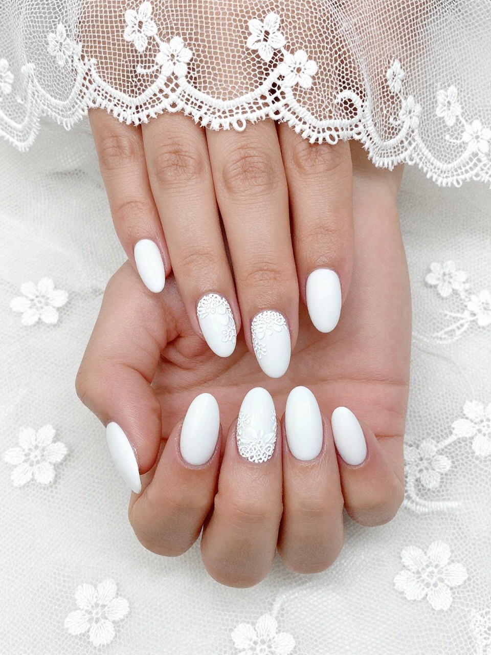 Bird's-eye view of hands with long, oval nails resting on a delicate lace bridal veil. The nails are an opaque matte white. An elaborate, textured white lace pattern with small floral shapes covers each nail. The lighting is bright and clean, highlighting the texture.