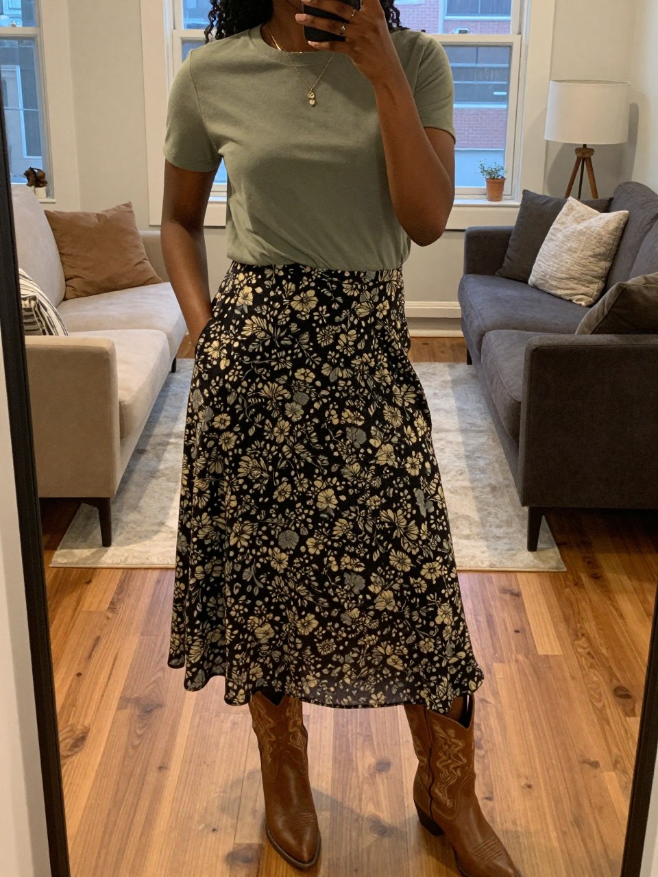 mirror selfie of a black woman in a sage green t-shirt and a floral skirt with western boots, one hand in her pocket, cropped at NECK so face doesn't show, in a city apartment, warm moody lighting, iPhone photo quality.