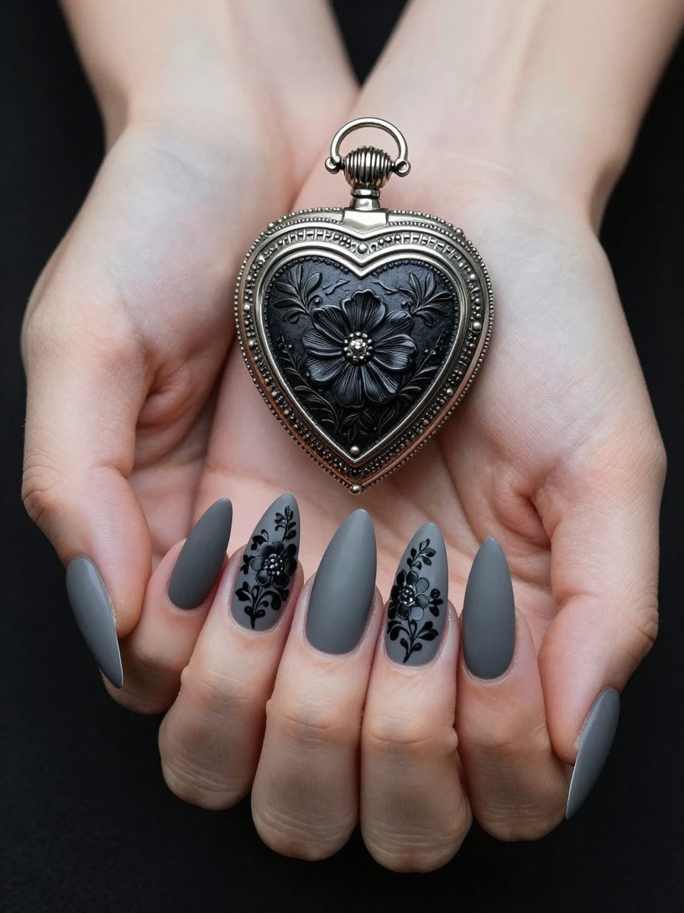 Overhead shot of hands with long, tapered nails resting on a vintage brooch. The nails are a matte charcoal grey. An ornate black and silver locket shape is painted on each nail, filled with micro black floral detailing. The lighting is dramatic.