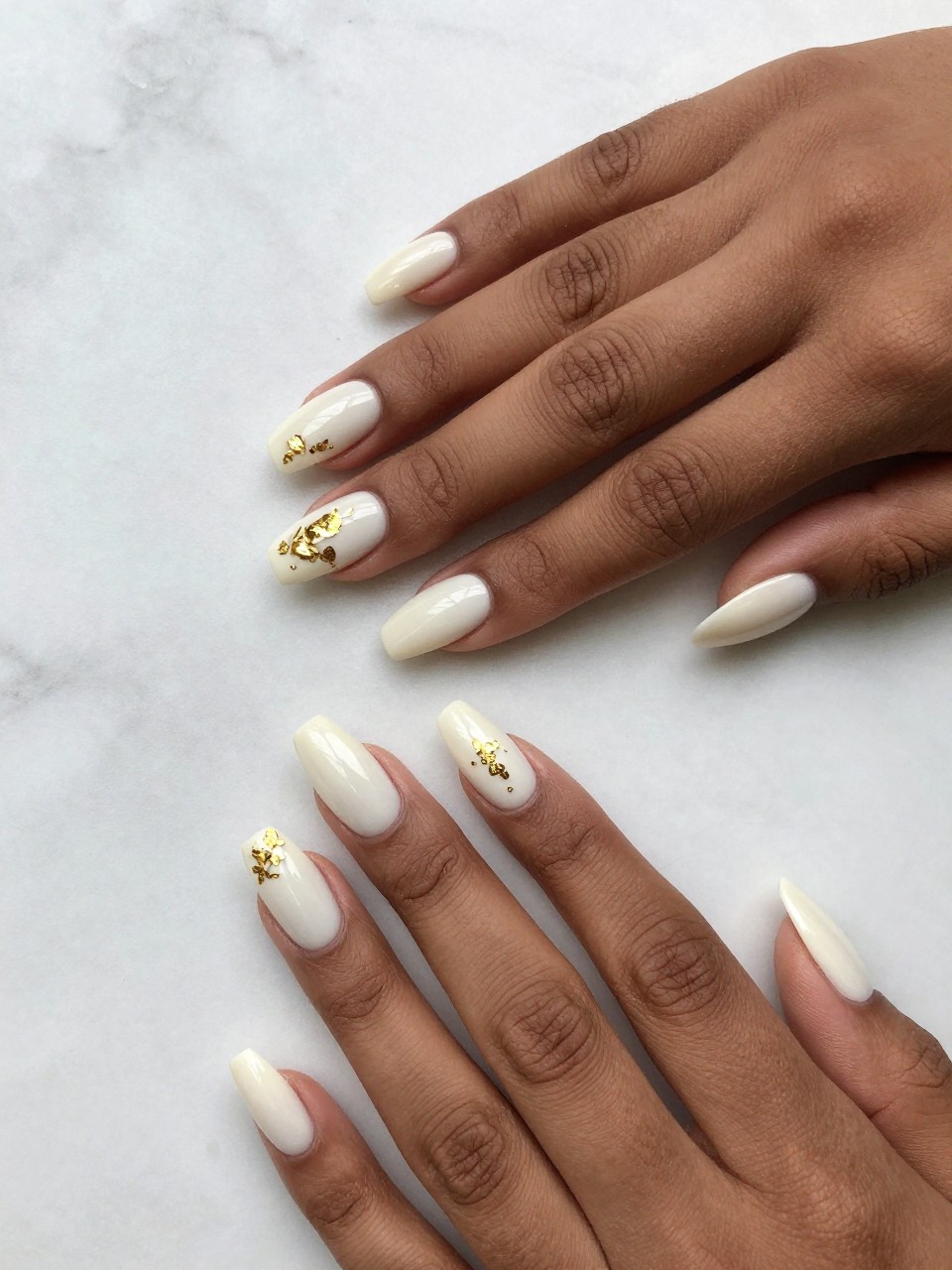 Top-down view of black feminine hands with coffin nails on a marble surface. The nails are a glossy ivory white. Tiny, fragmented pieces of gold leaf are arranged in small, loose clusters on each nail. The bright light makes the gold shimmer delicately.