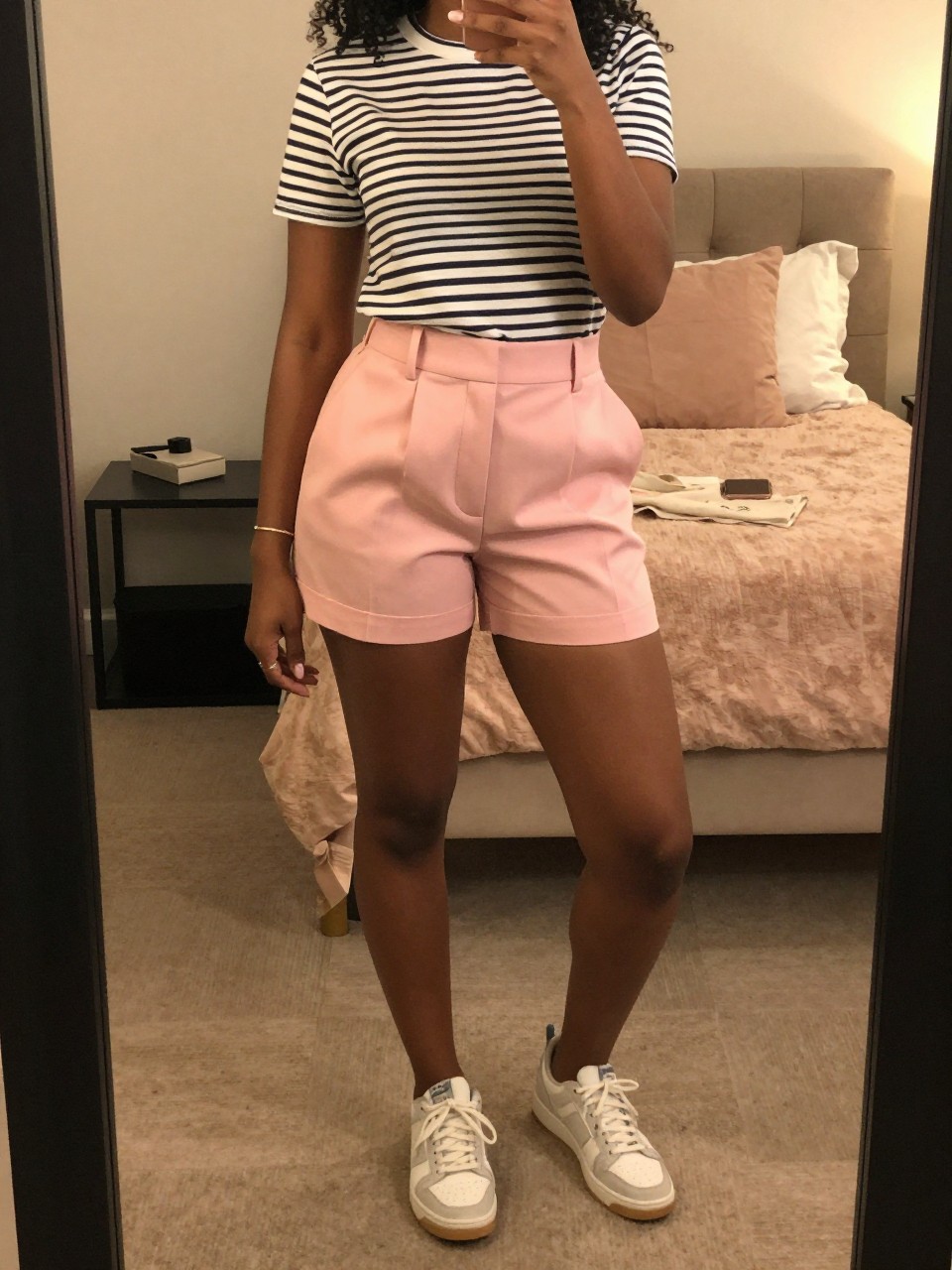 mirror selfie of a black woman in pink tailored shorts and a striped tee with sneakers, standing with weight shifted, cropped at NECK so face doesn't show, in a feminine-themed bedroom, warm moody lighting, iPhone photo quality.