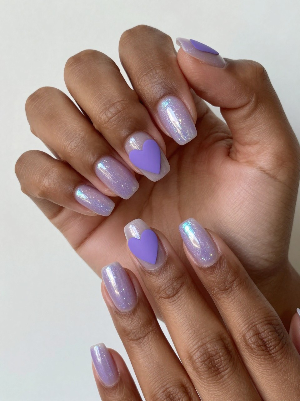Black feminine hands moving gently under a light to show holographic sparkle. The nails have a sheer holographic lavender base. A soft, matte purple heart is centered on each nail, absorbing light amidst the sparkle.