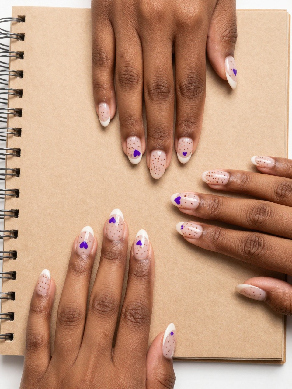 Overhead view of black feminine hands on a kraft paper journal. The nails have a sheer nude base with light brown freckles design and a few micro purple hearts are hidden on the nails too. The look is playful, organic, and sweet.