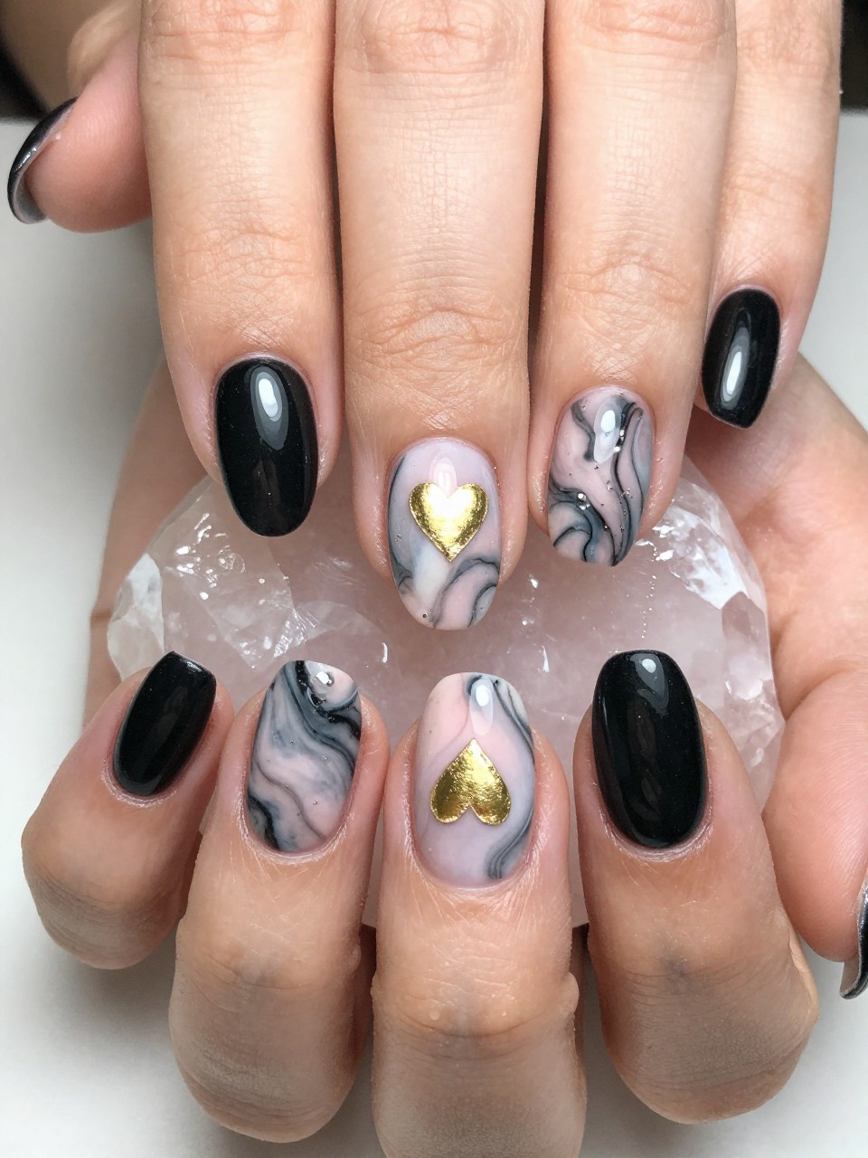 Close-up, top-down view of a black feminine nail with a pink and white marble swirl. A single, small gold foil heart is embedded in the design. The hands rest on a rose quartz crystal, complementing the elegant vibe.