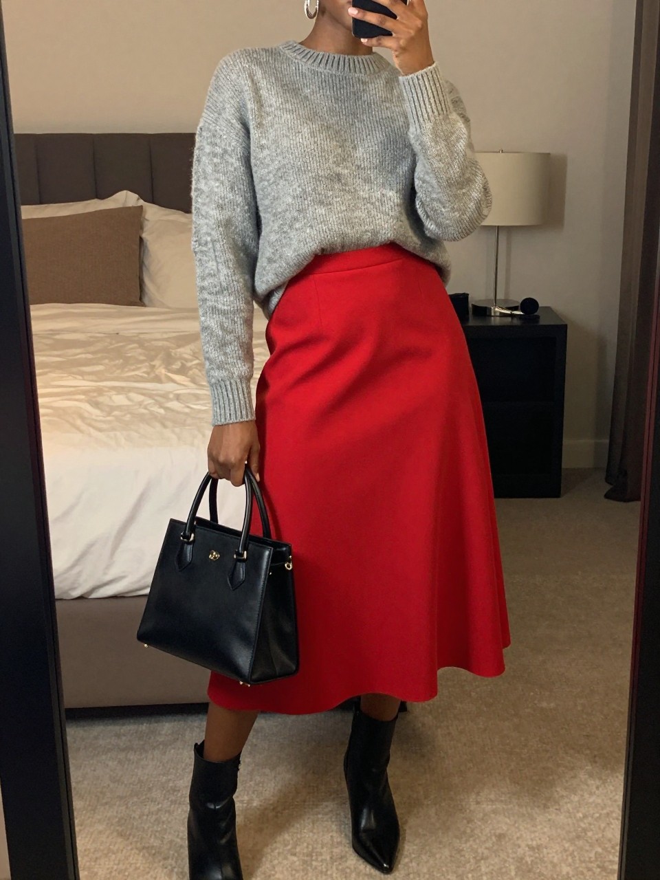 mirror selfie of a black woman wearing a heather gray fine-knit sweater tucked into a bright red A-line midi skirt with black ankle boots and silver hoop earrings, holding a black structured bag, standing in an elegant pose, cropped at NECK so face doesn't show, in a feminine-themed bedroom, warm moody lighting, iPhone photo quality.