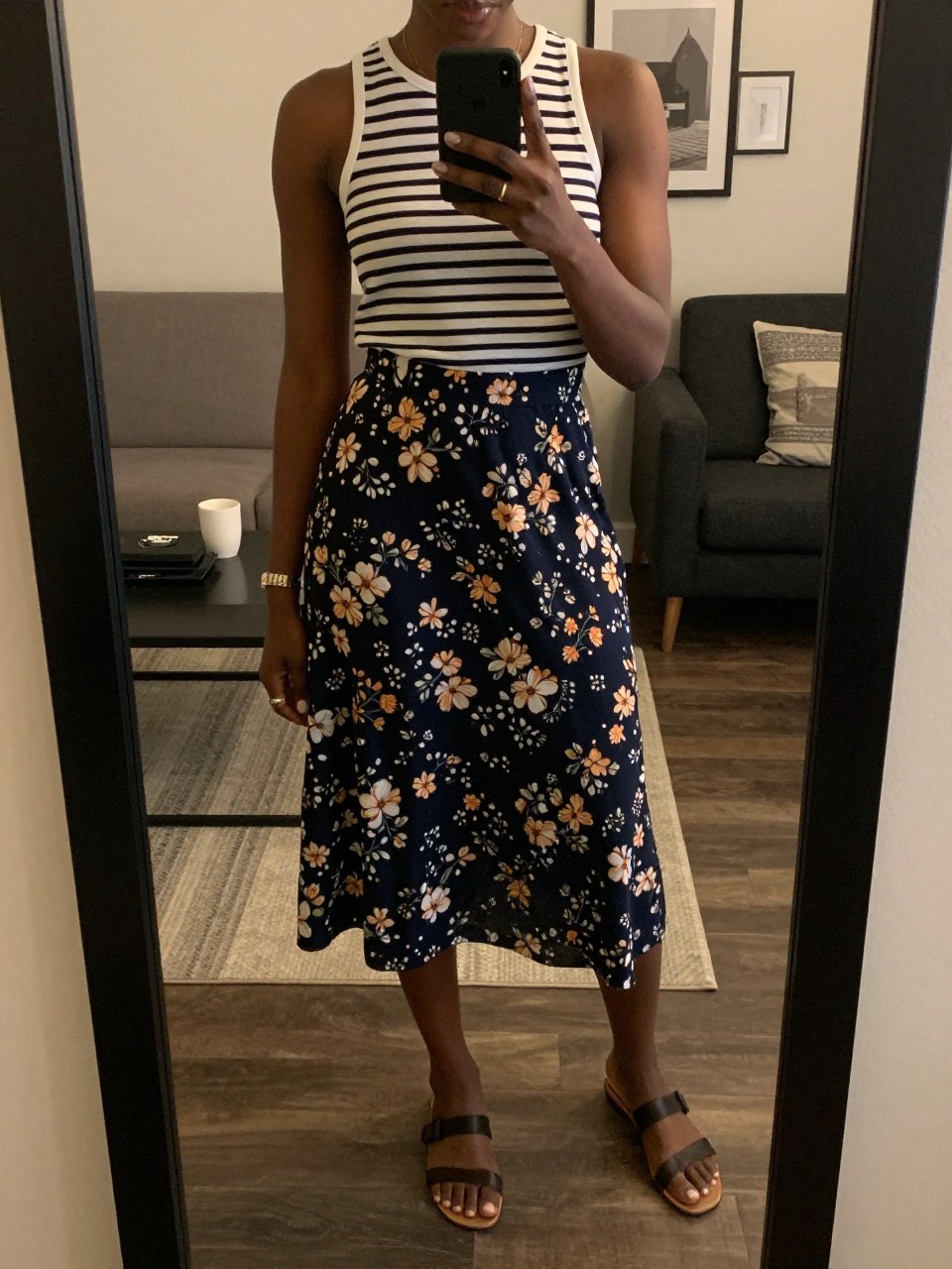iPhone mirror selfie of a black woman in a striped top and a navy floral skirt with flat sandals, casually holding her phone at her side, cropped at NECK so face doesn't show, in a city apartment, warm moody lighting, iPhone photo quality.